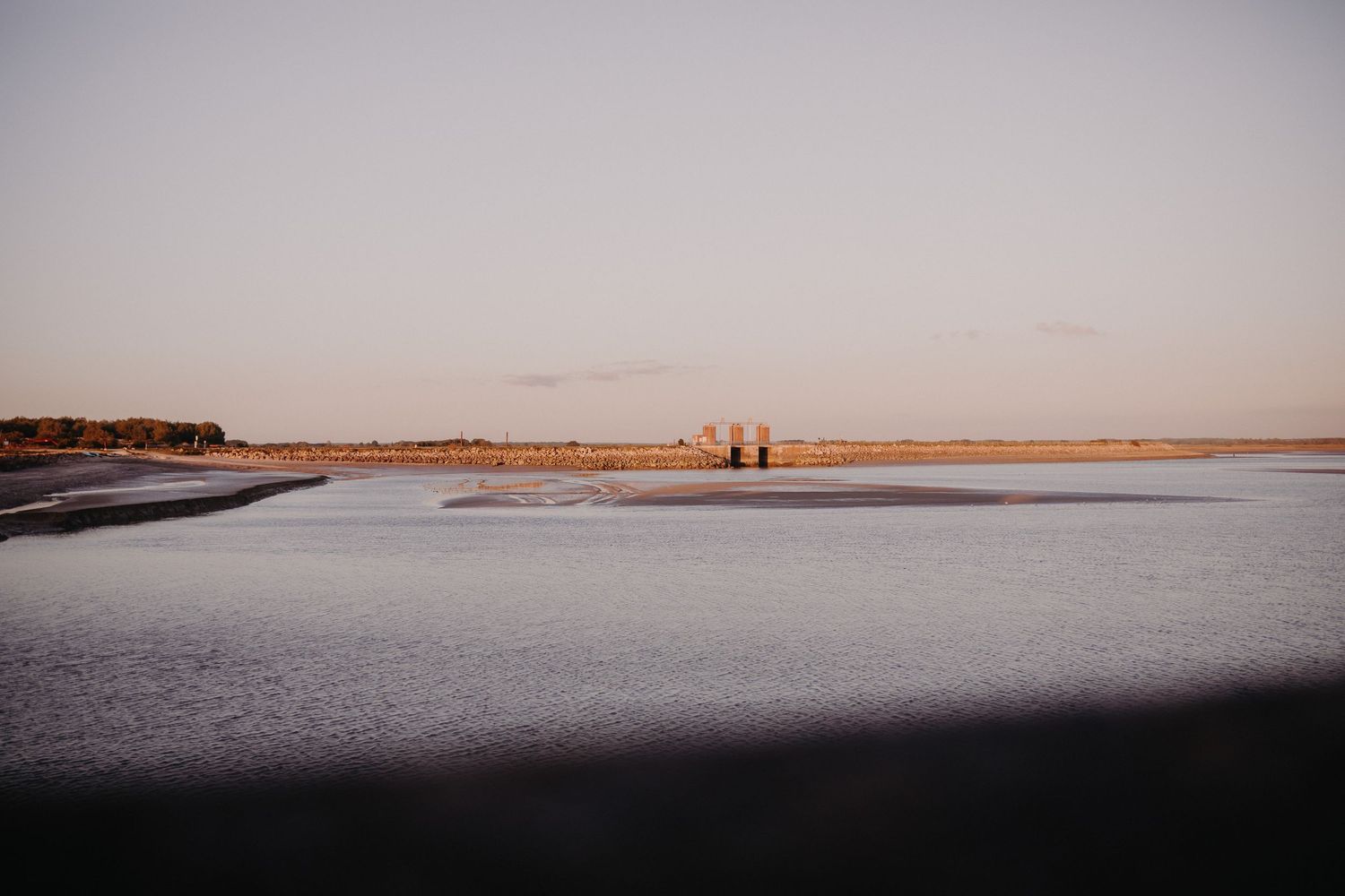 Two silhouettes walk along a tranquil beach at sunset with a sandy shore and calm waters stretching to the horizon.