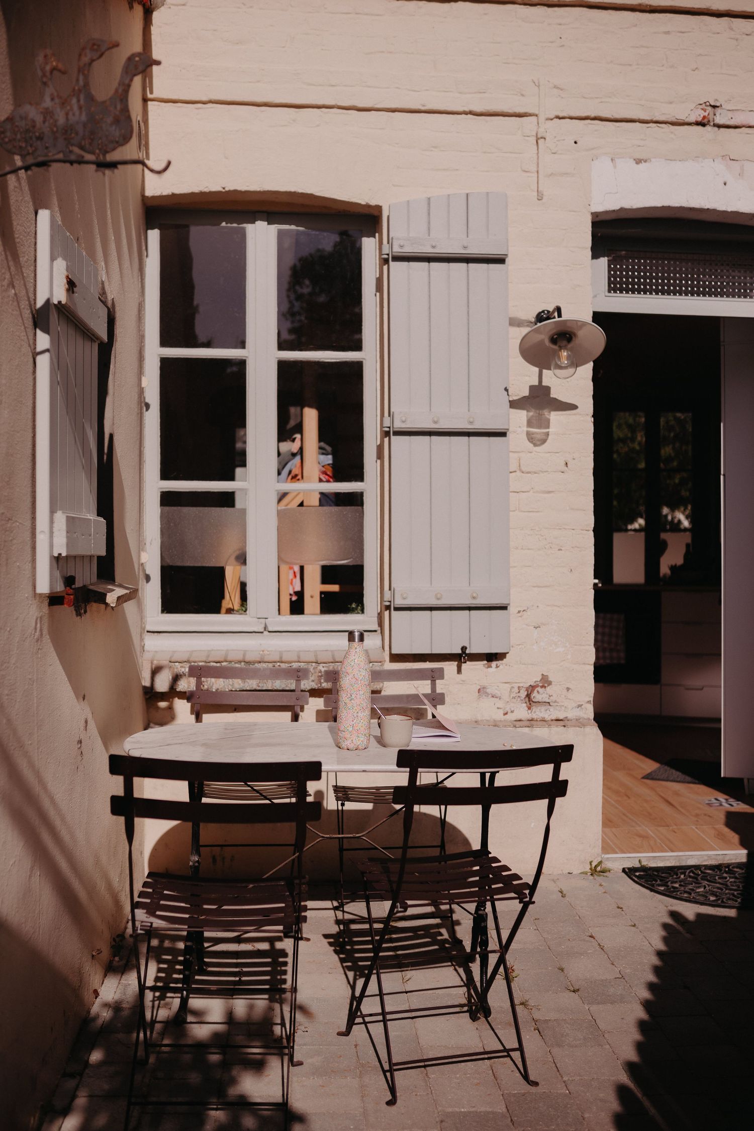 Rustic French cafe terrace with folding black chairs and tables in front of a white building with shuttered windows.