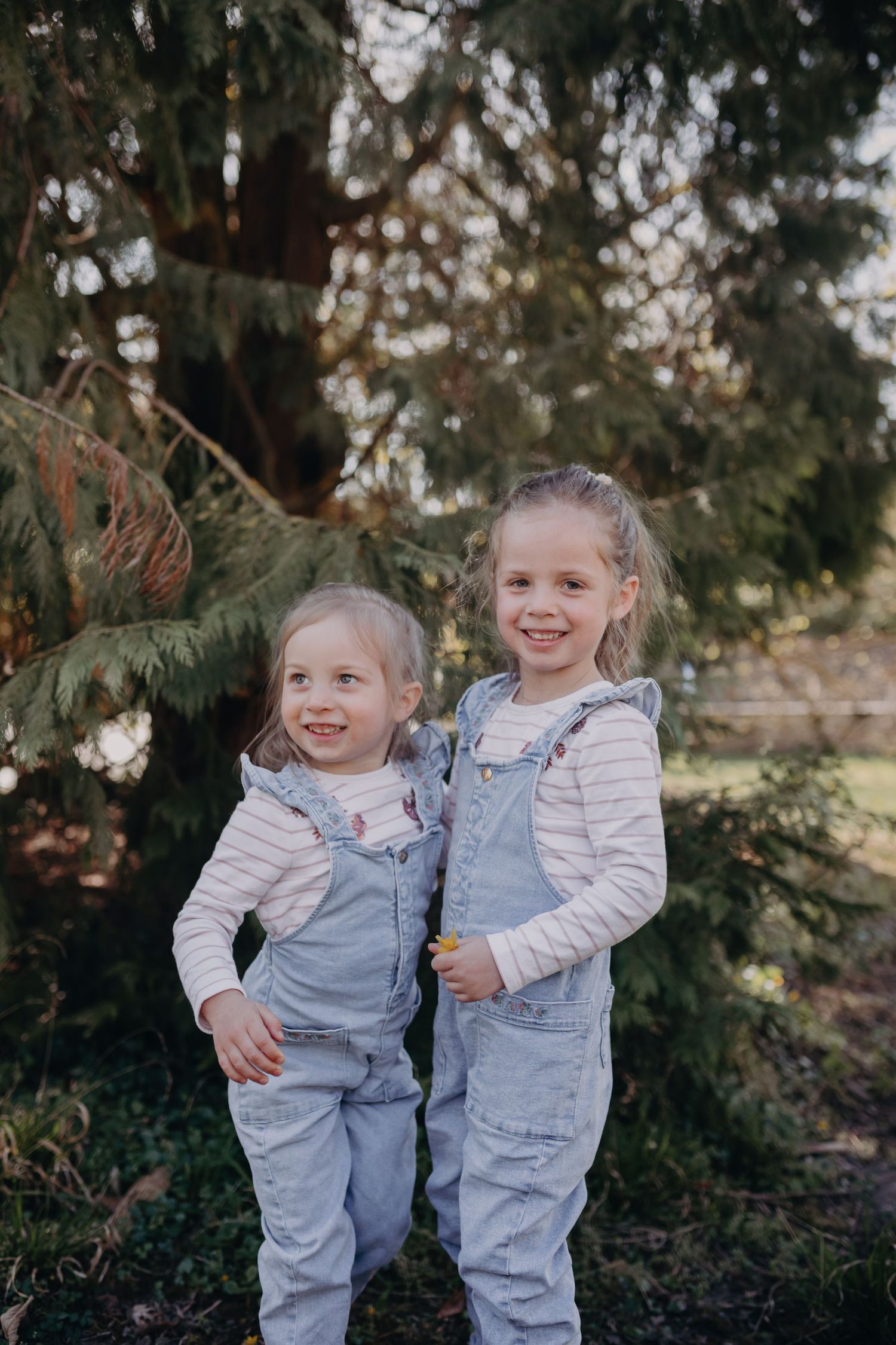 Two small children wearing matching gray overalls and white long sleeve shirts stand together near evergreen trees.
