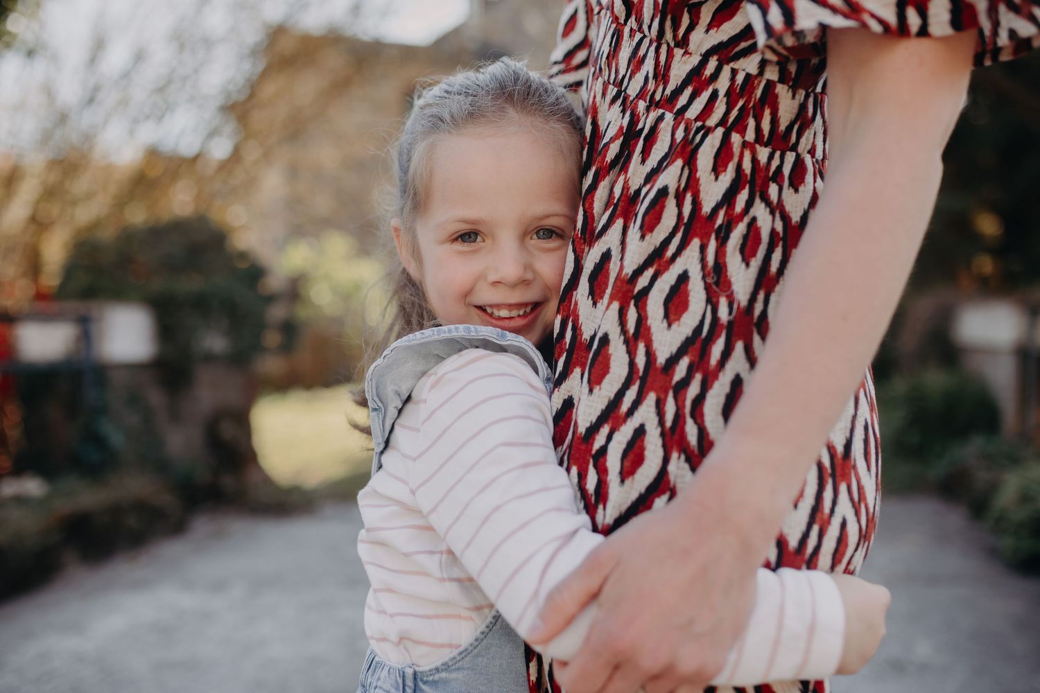 Child in pink sweater hugs adult wearing patterned dress outdoors on sunny day.