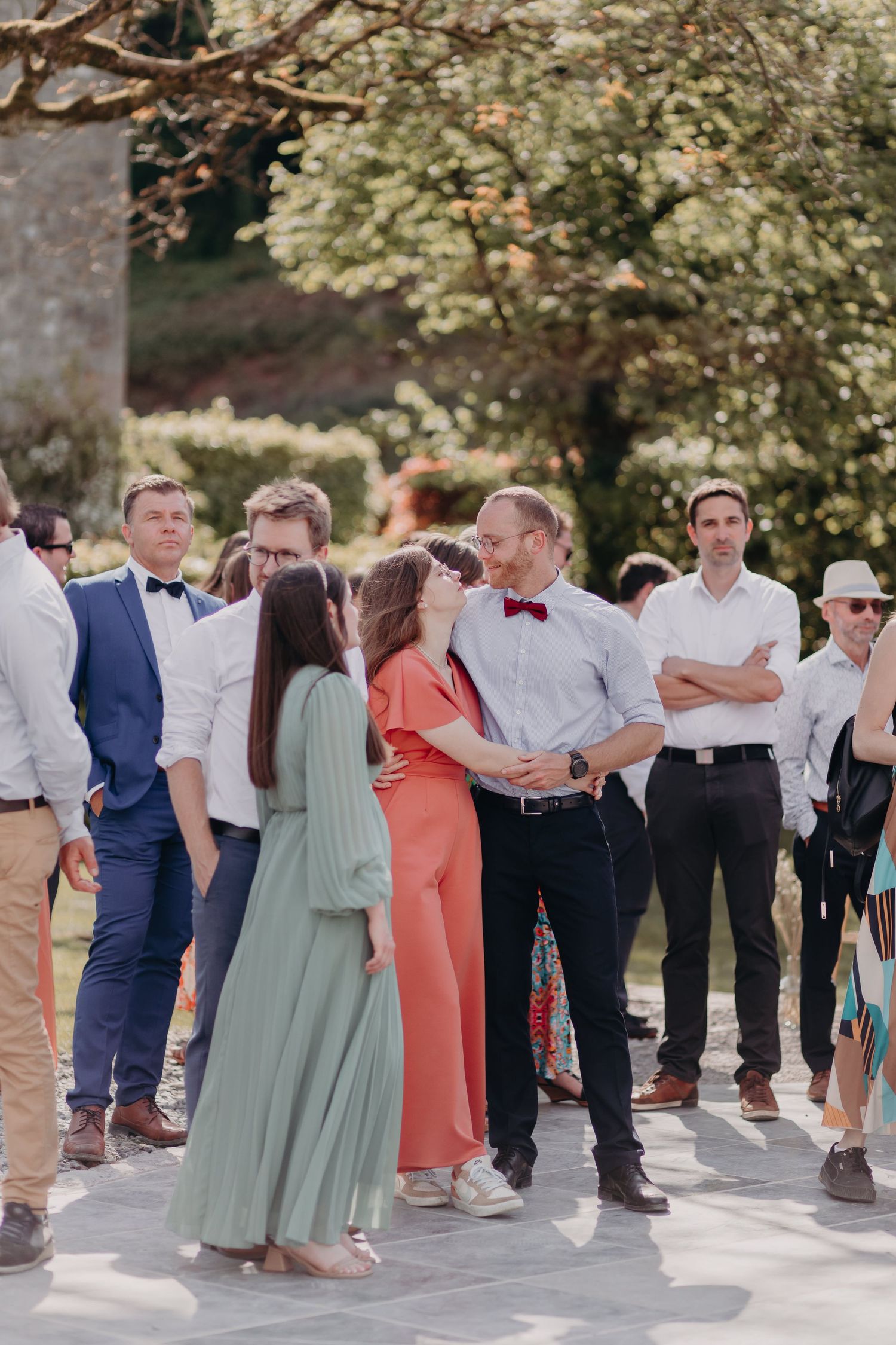 A group of wedding guests in formal attire socialize outdoors under trees during a summer celebration.