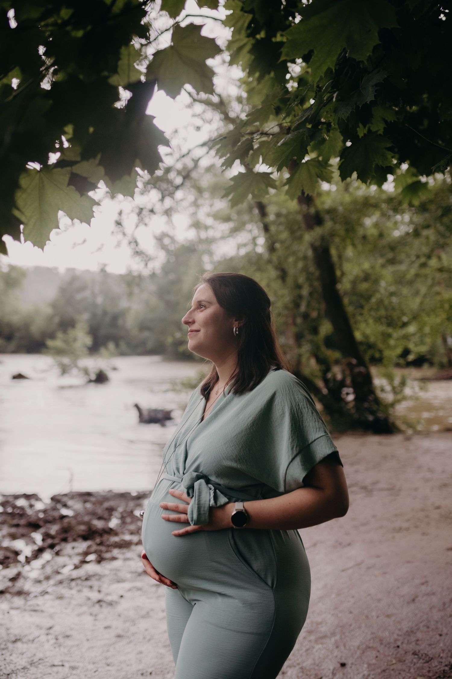 Pregnant woman in sage green dress stands peacefully by a river under tree branches on misty morning.