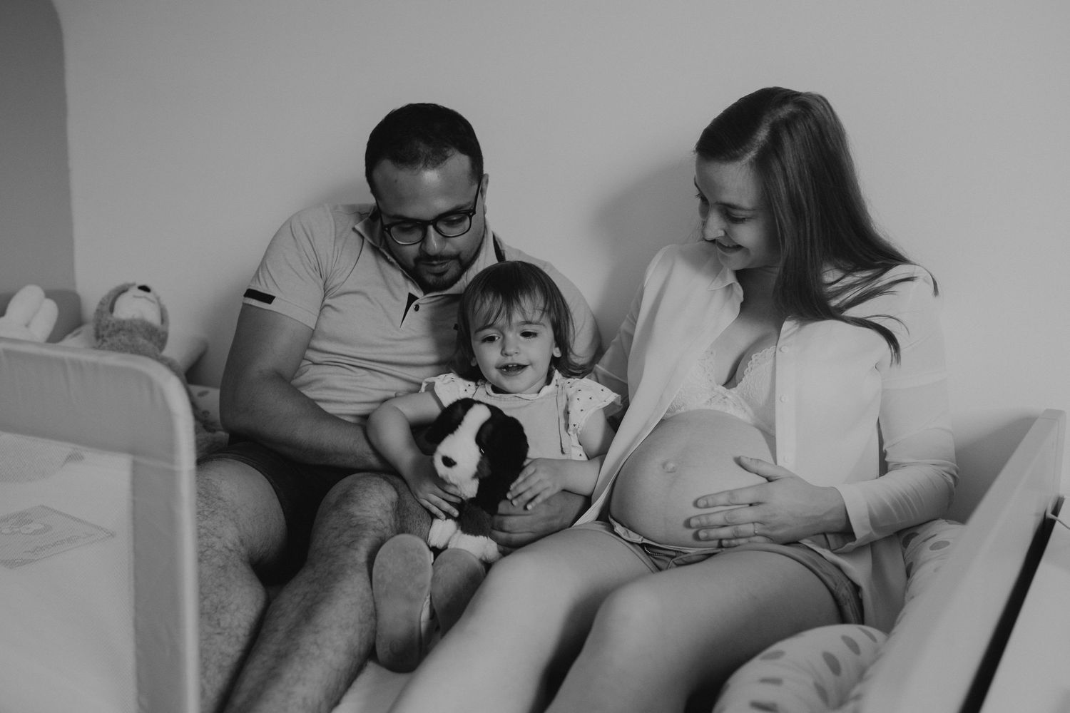 A black and white photo shows a family resting together on a couch with their dog while expecting a new addition.