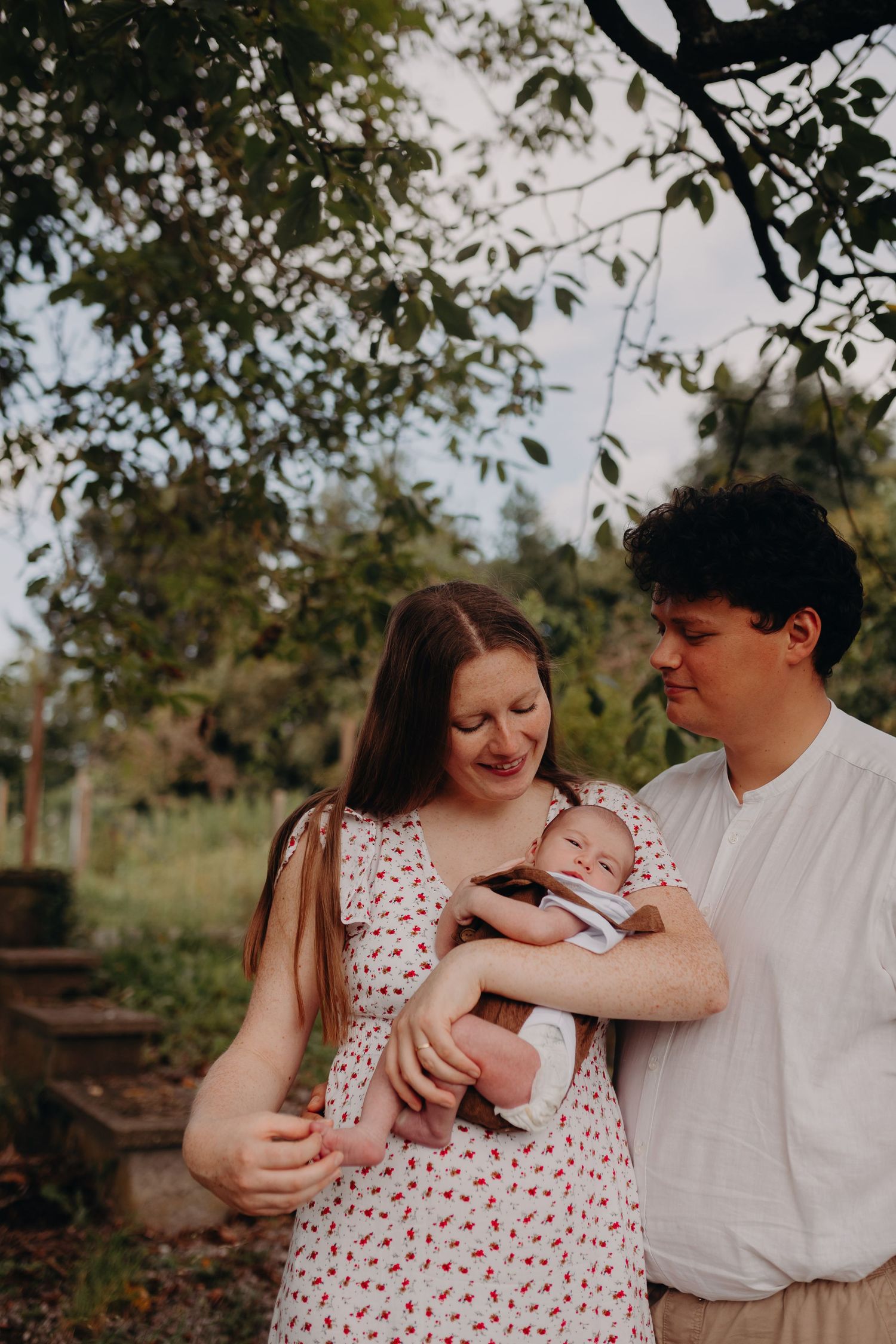 A couple stands together outdoors holding their baby under tree branches on a sunny day.