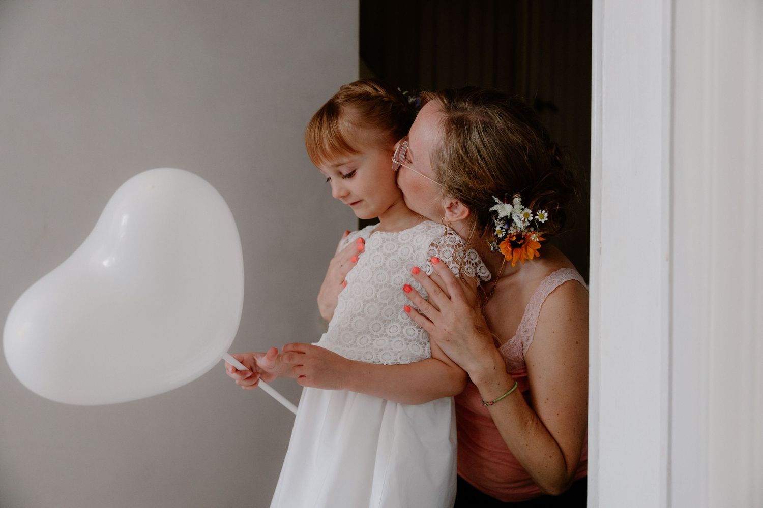 A young flower girl holds a white heart balloon while a woman in pink kisses her cheek at a wedding.