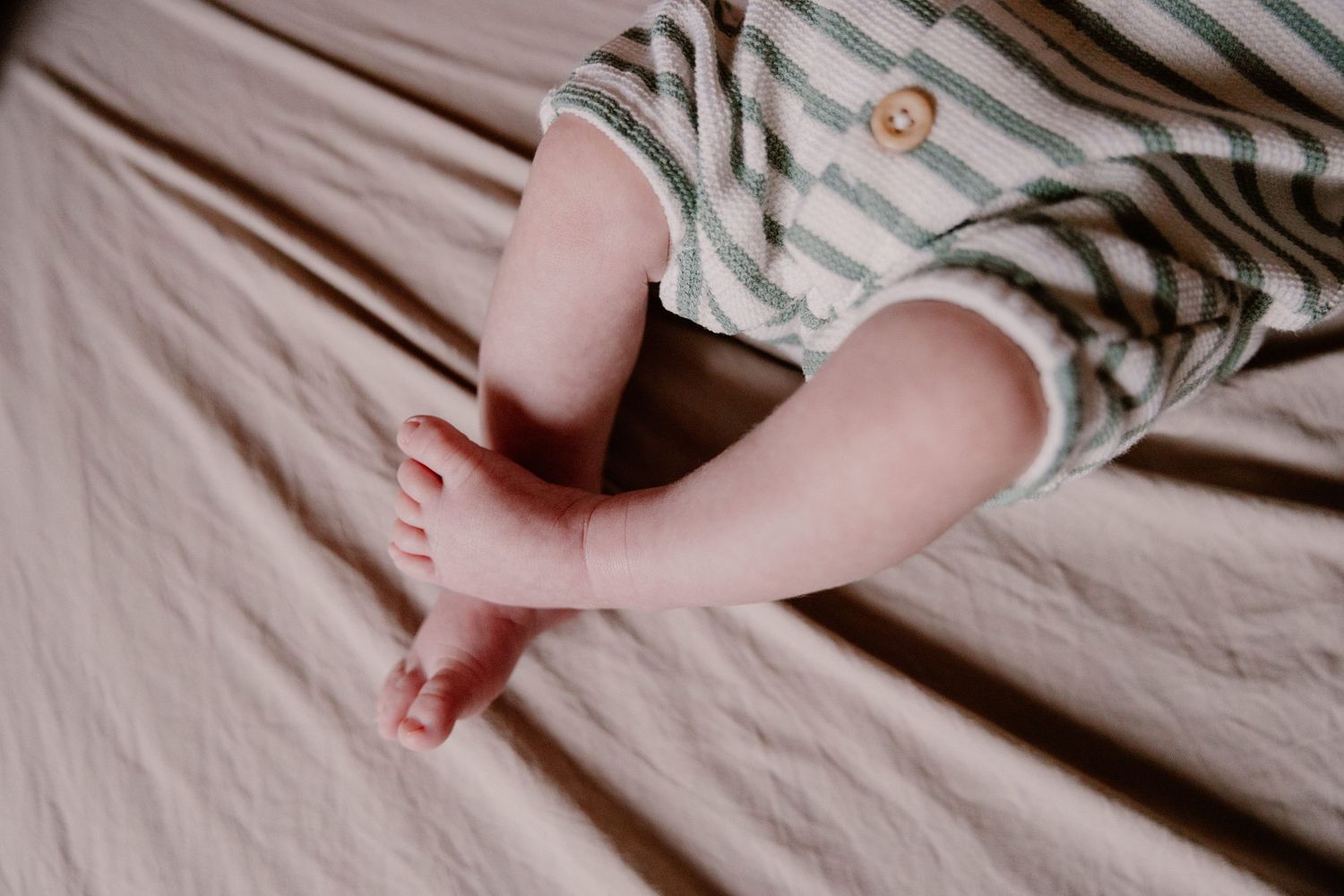 Close-up of a baby's tiny feet and legs in a striped green and white romper lying on a soft pink linen surface.
