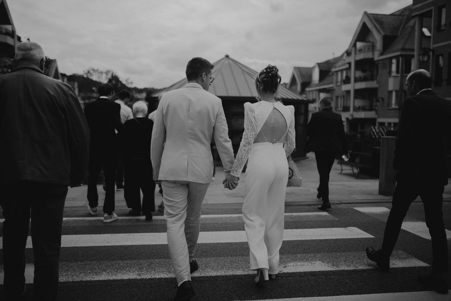 A couple dressed in white holding hands walking together on a street among other pedestrians in a black and white photo.