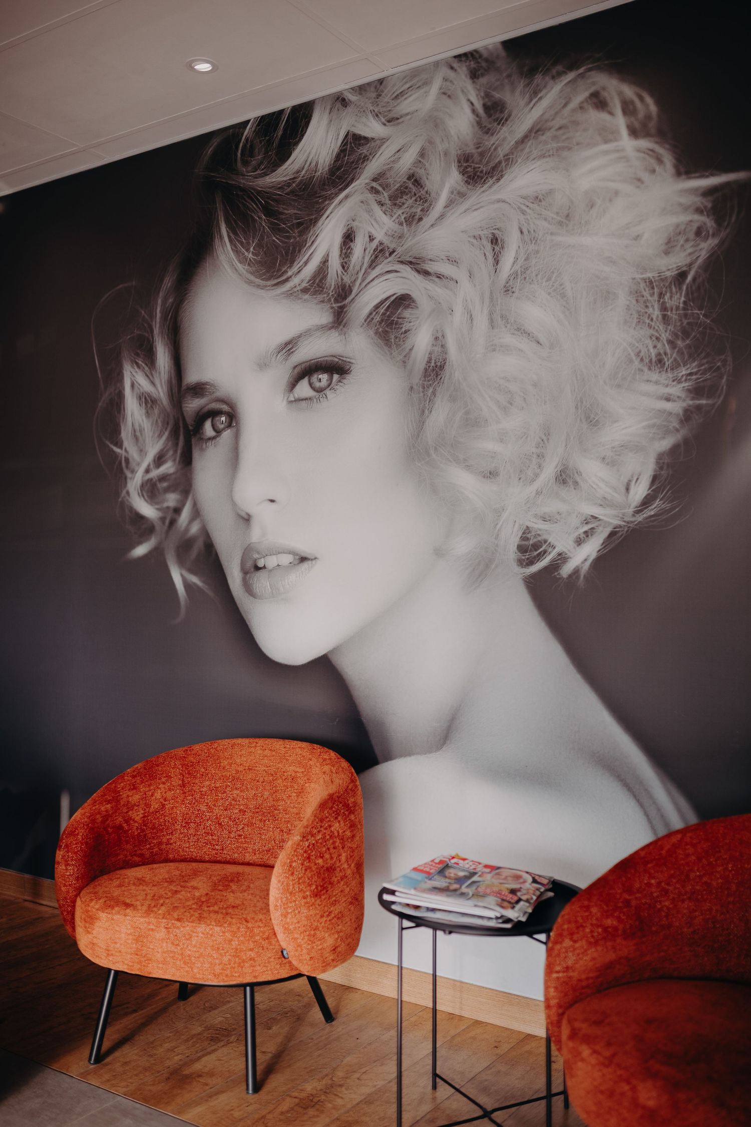 Modern salon interior with large black and white photograph on wall and two orange accent chairs flanking small table.