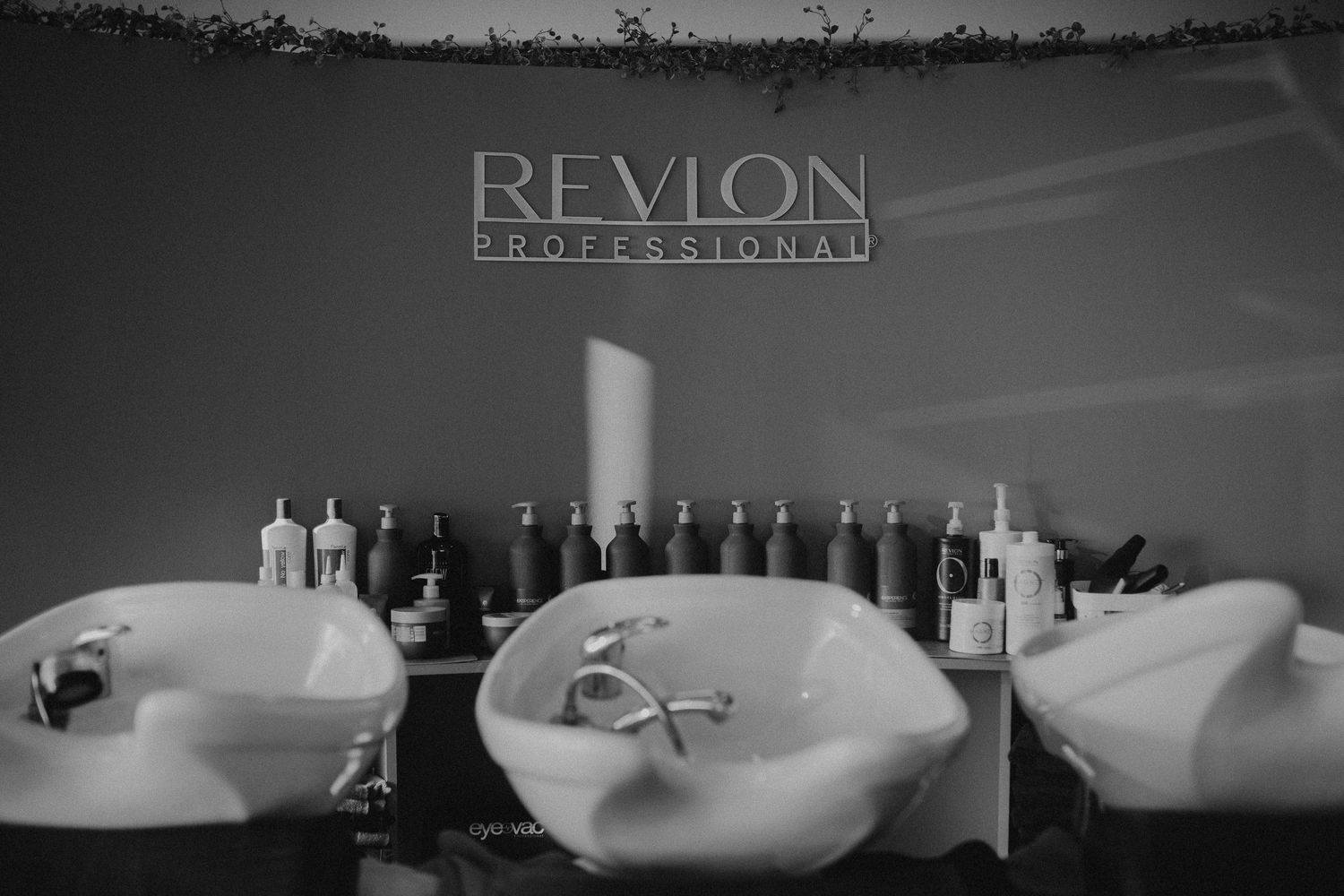 Black and white photo of salon hair washing stations with Revlon Professional branding and lined-up haircare products.