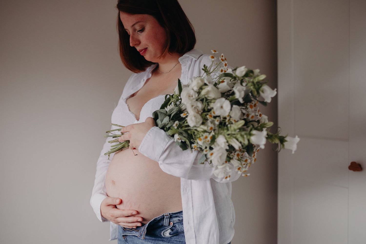 A pregnant woman in a white blouse holds an elegant bouquet of white flowers against a neutral background.