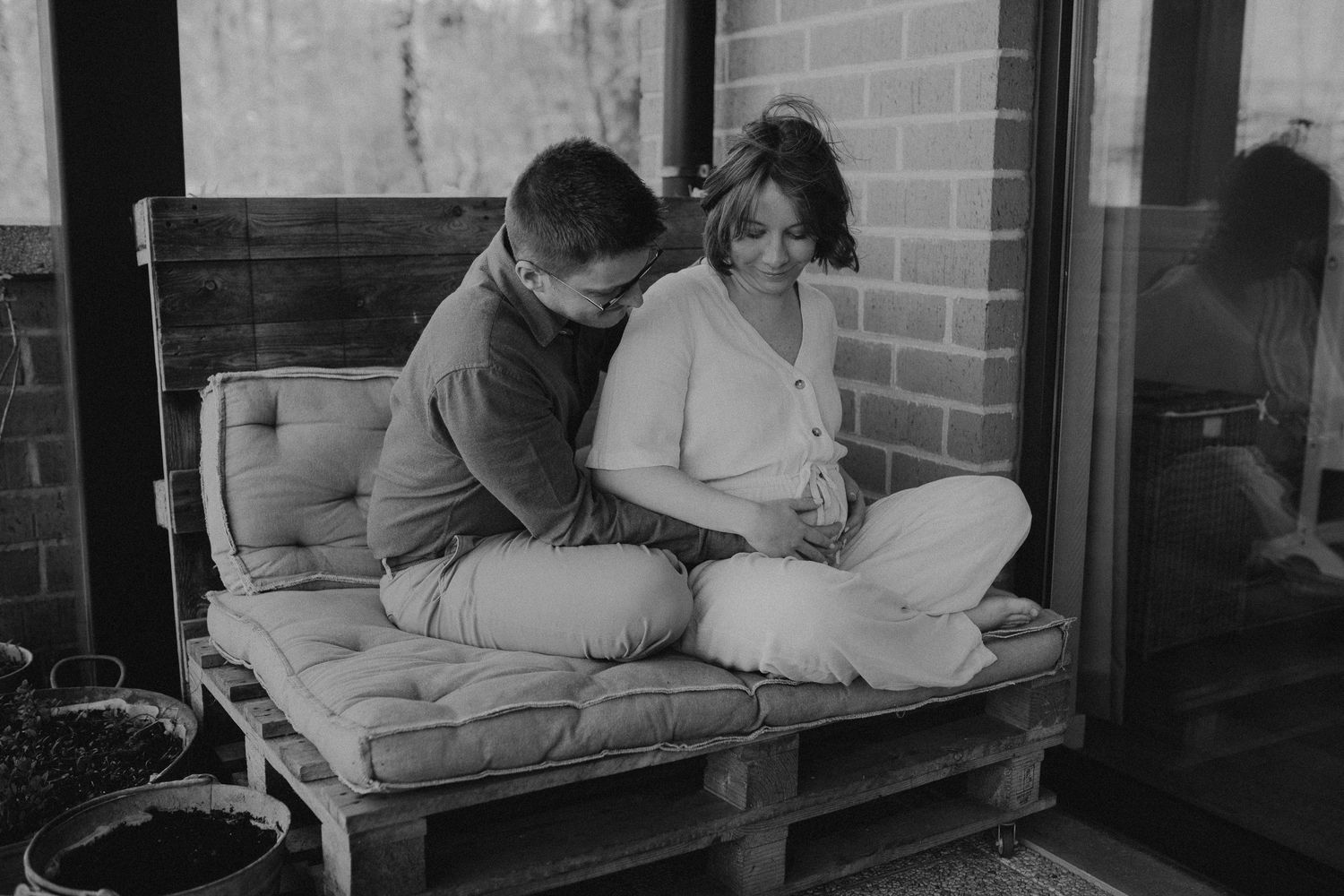 Black and white photo of two people sitting together on cushions on a rustic porch setting.