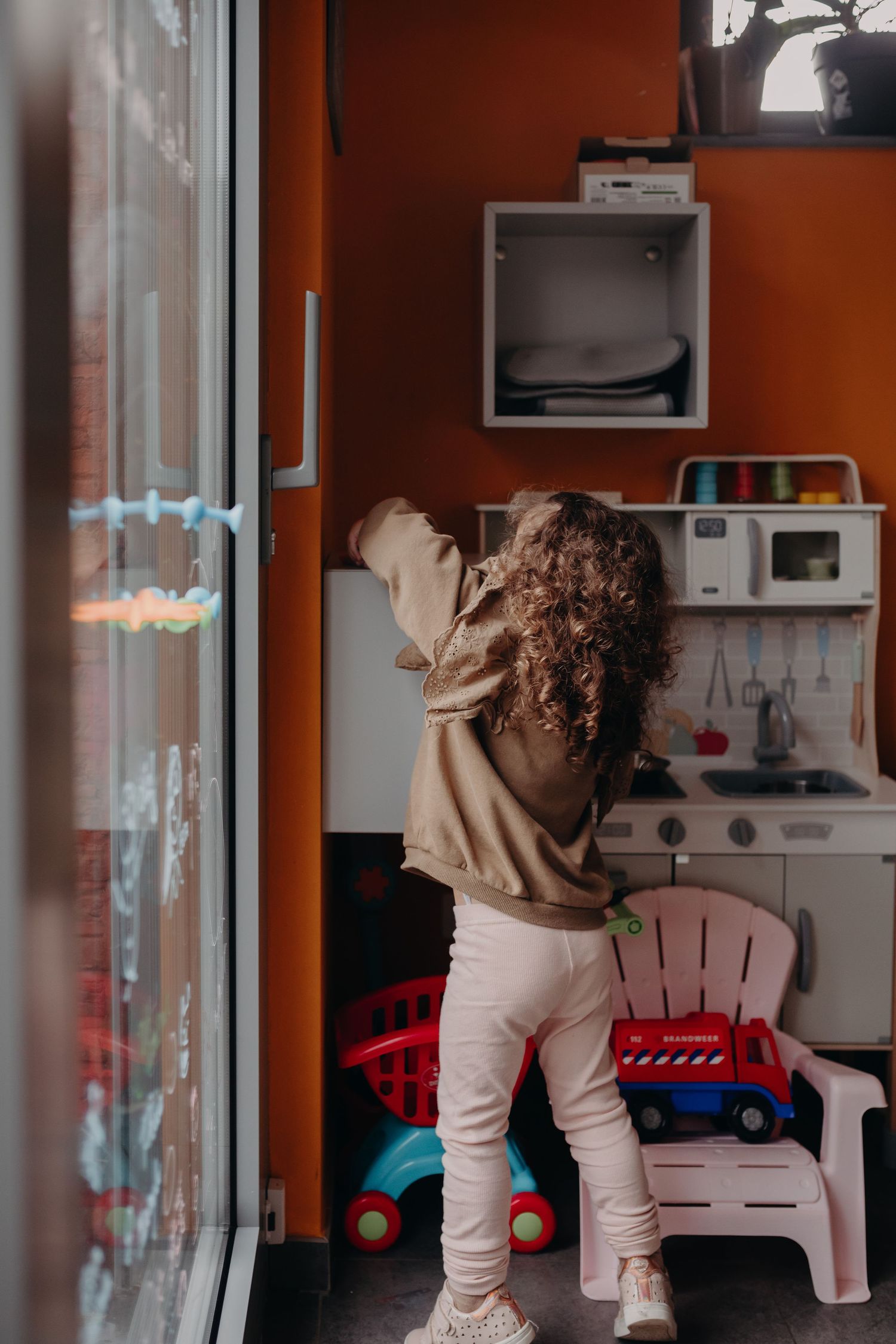 A small child in beige sweater and white pants plays in a toy kitchen area near a window.