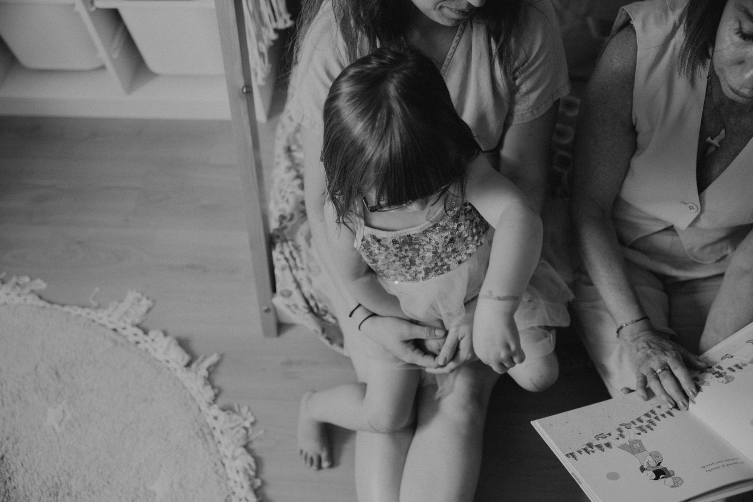 Black and white photo of a young girl sitting on floor reading a book with family members nearby.
