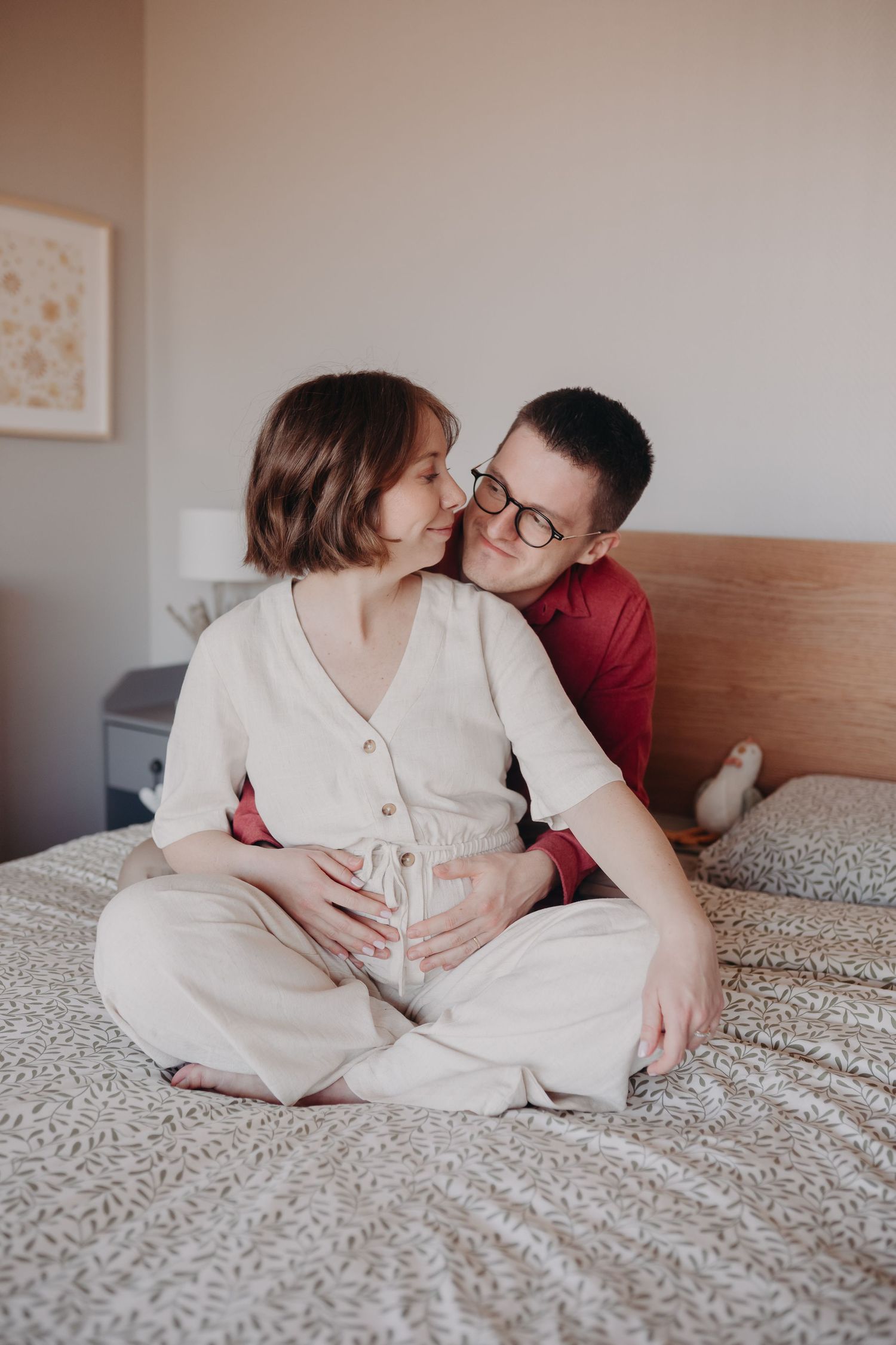 Affectionate couple sits on gray bedding while sharing an intimate moment at home in soft lighting.