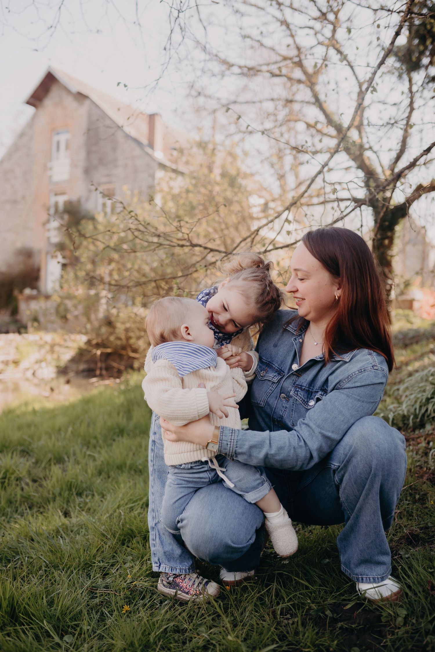 A mother in denim kneels on grassy ground with a baby in front of a rustic house at golden hour.