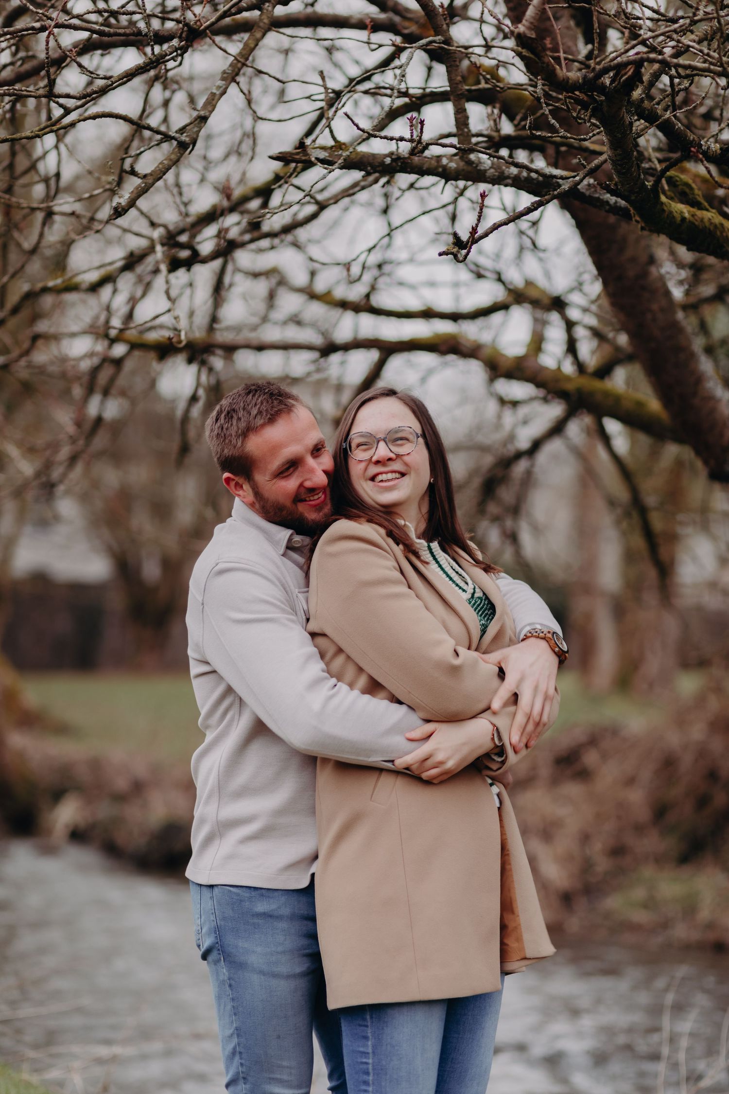 A couple shares a joyful embrace while standing on a path surrounded by bare winter trees.