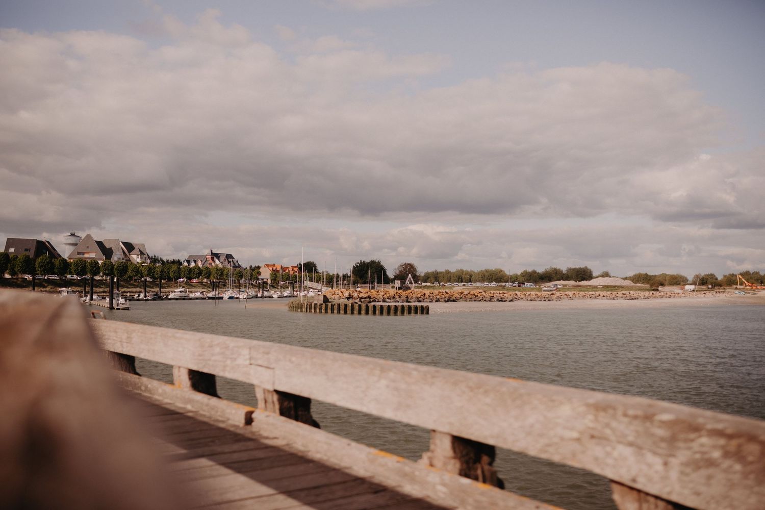 Scenic coastal view of a concrete seawall overlooking a marina with boats and beachfront buildings under cloudy skies.