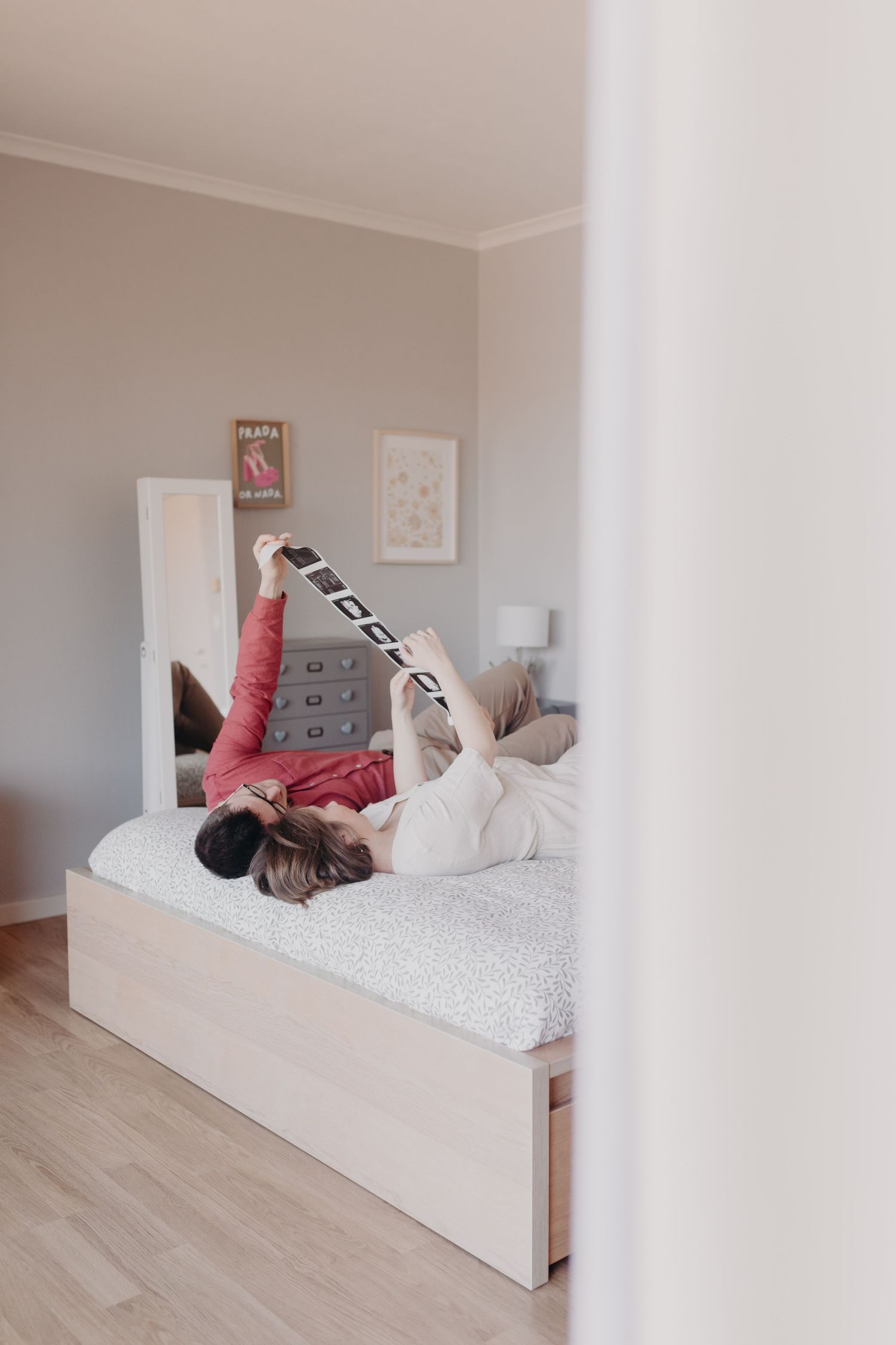 Someone relaxes on a white bed with a smartphone in a minimalist bedroom with light gray walls and wooden flooring.