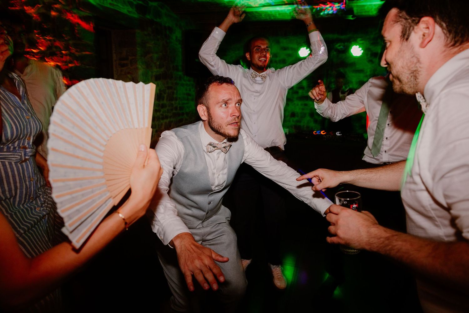 Guests dancing energetically at a wedding reception party with green lighting in a rustic venue.