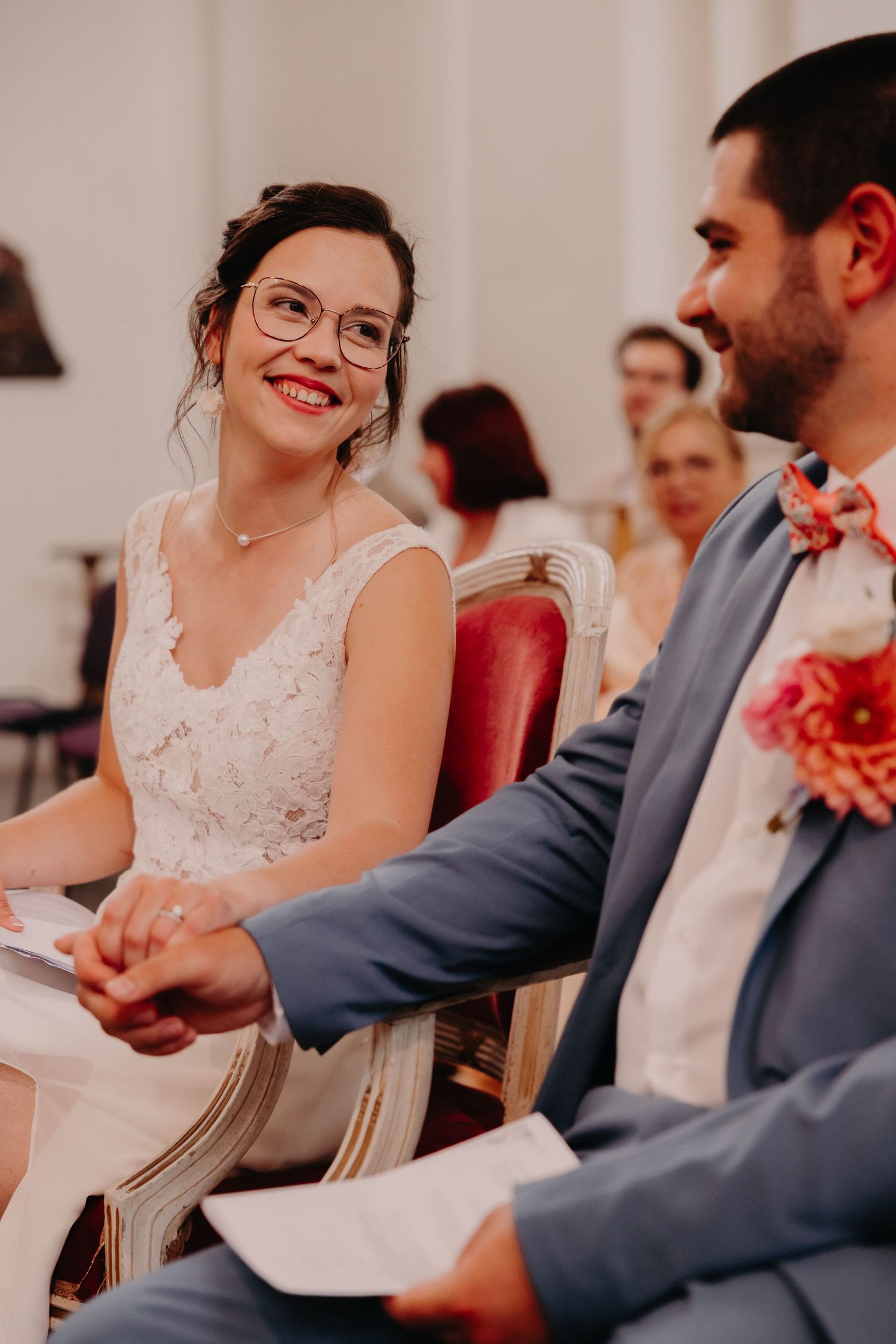 Wedding ceremony attendees smile and laugh while seated in elegant red chairs during an indoor celebration.