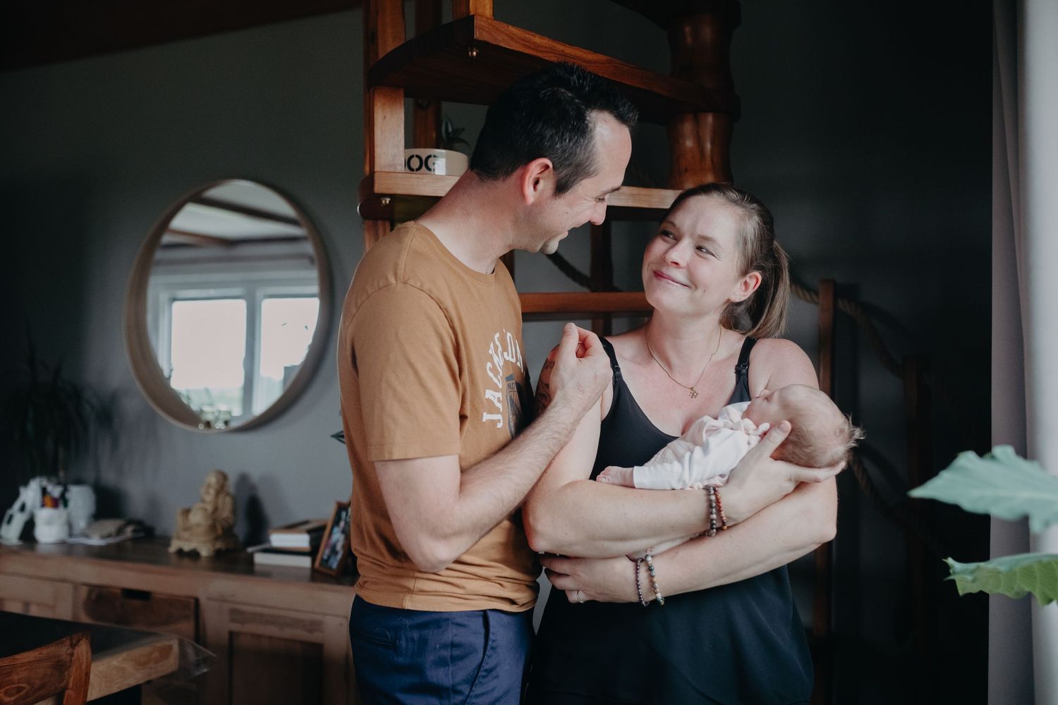 Two people share an intimate moment while holding a small infant in a cozy home setting with a circular window.