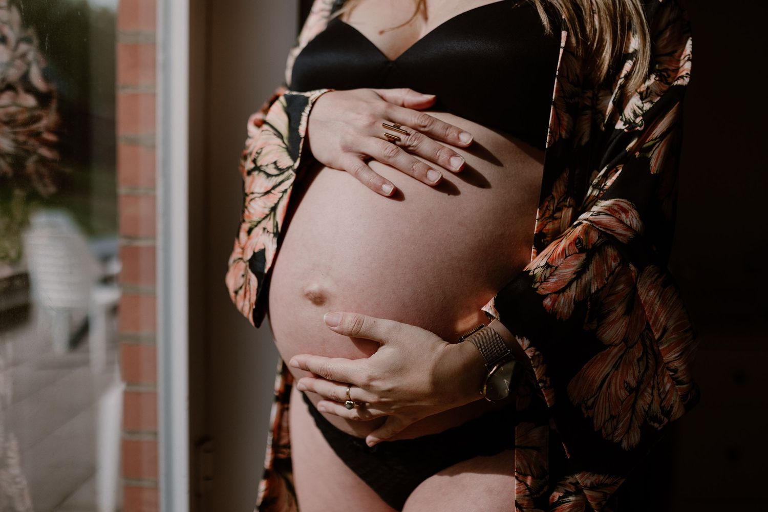 Close up maternity photo showing pregnant belly with hands gently cradling baby bump while wearing black lingerie and floral robe.