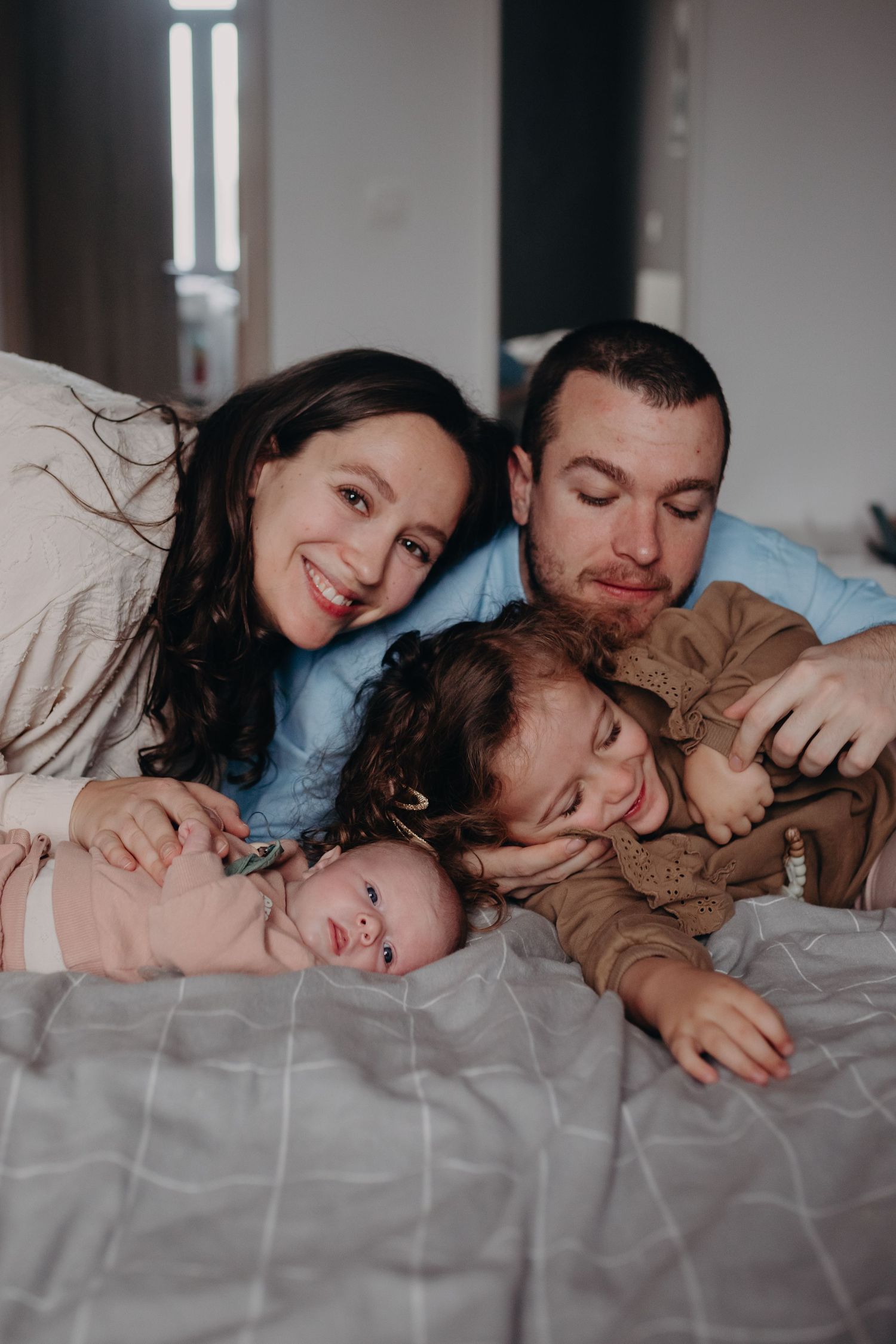 A family of four cuddles together on gray bedding during an intimate lifestyle photography session at home.