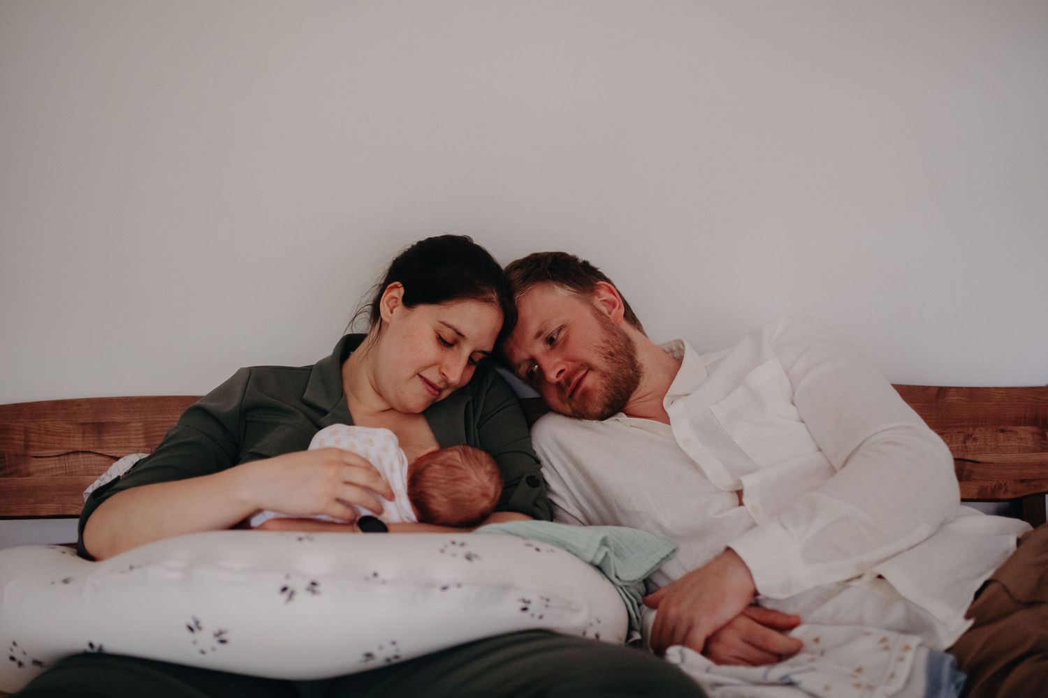 Family rests together on a wooden headboard while cuddling their newborn baby at home.