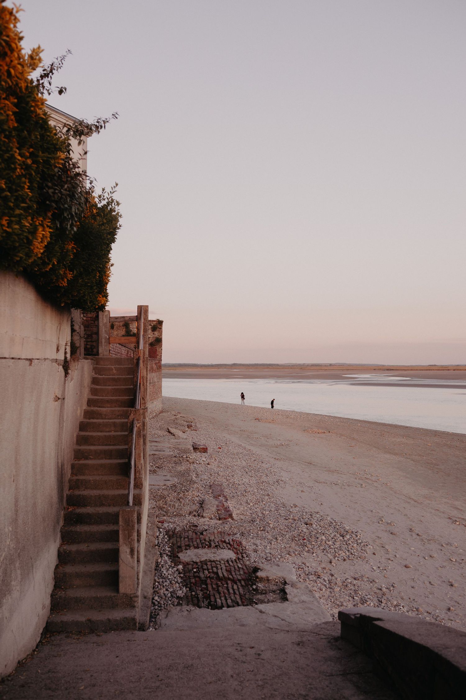 Concrete stairs lead down to a secluded beach at sunset with distant figures walking along the shore.