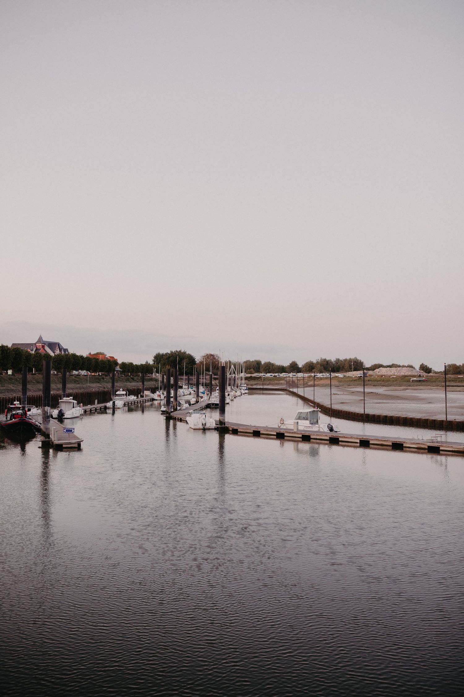Boats moored at a tranquil marina during dusk with industrial buildings and trees lining the waterfront.