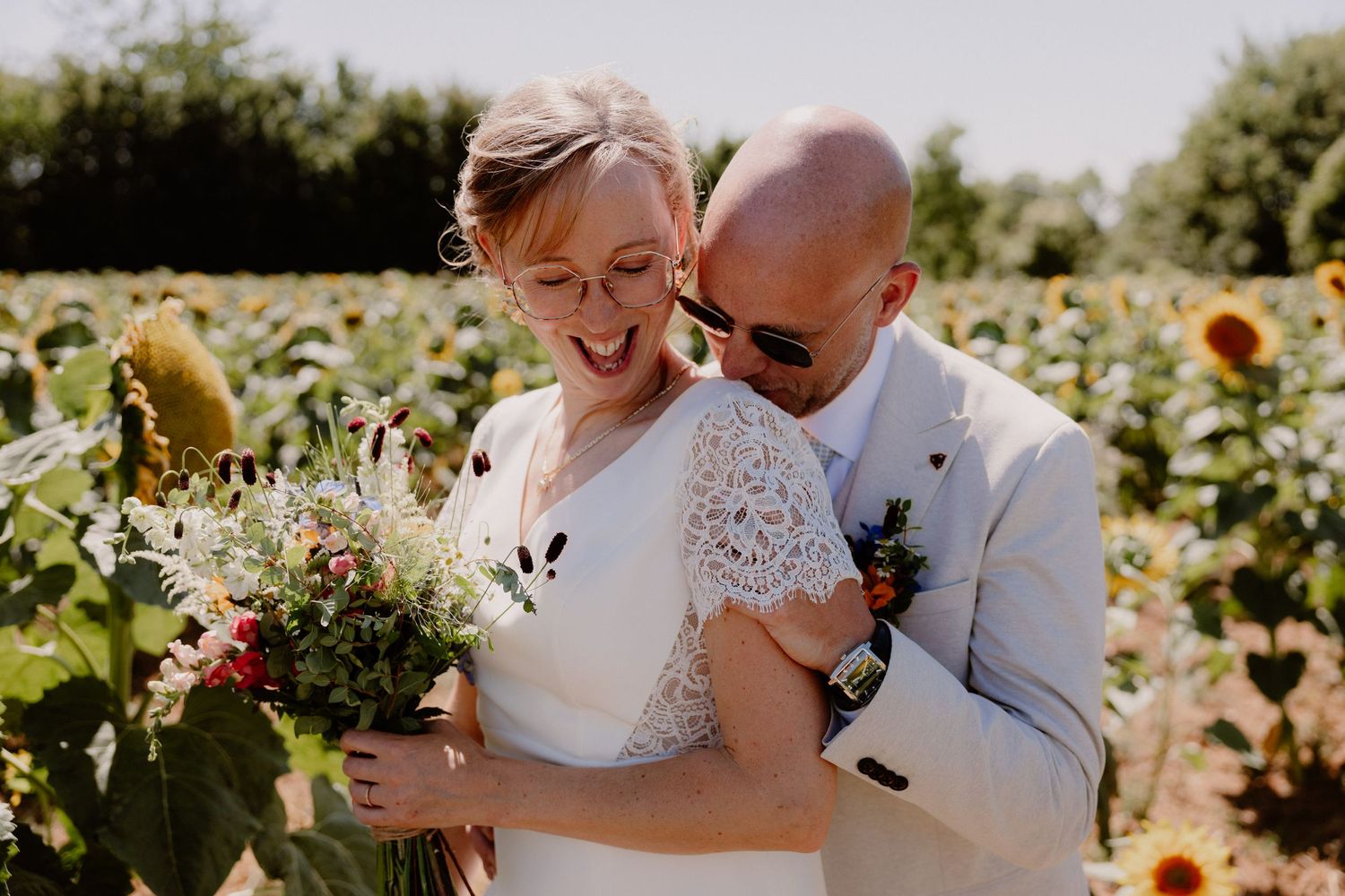 Bride and groom embracing in a sunflower field, bride holding a colorful bouquet, both smiling joyfully on a sunny wedding day.