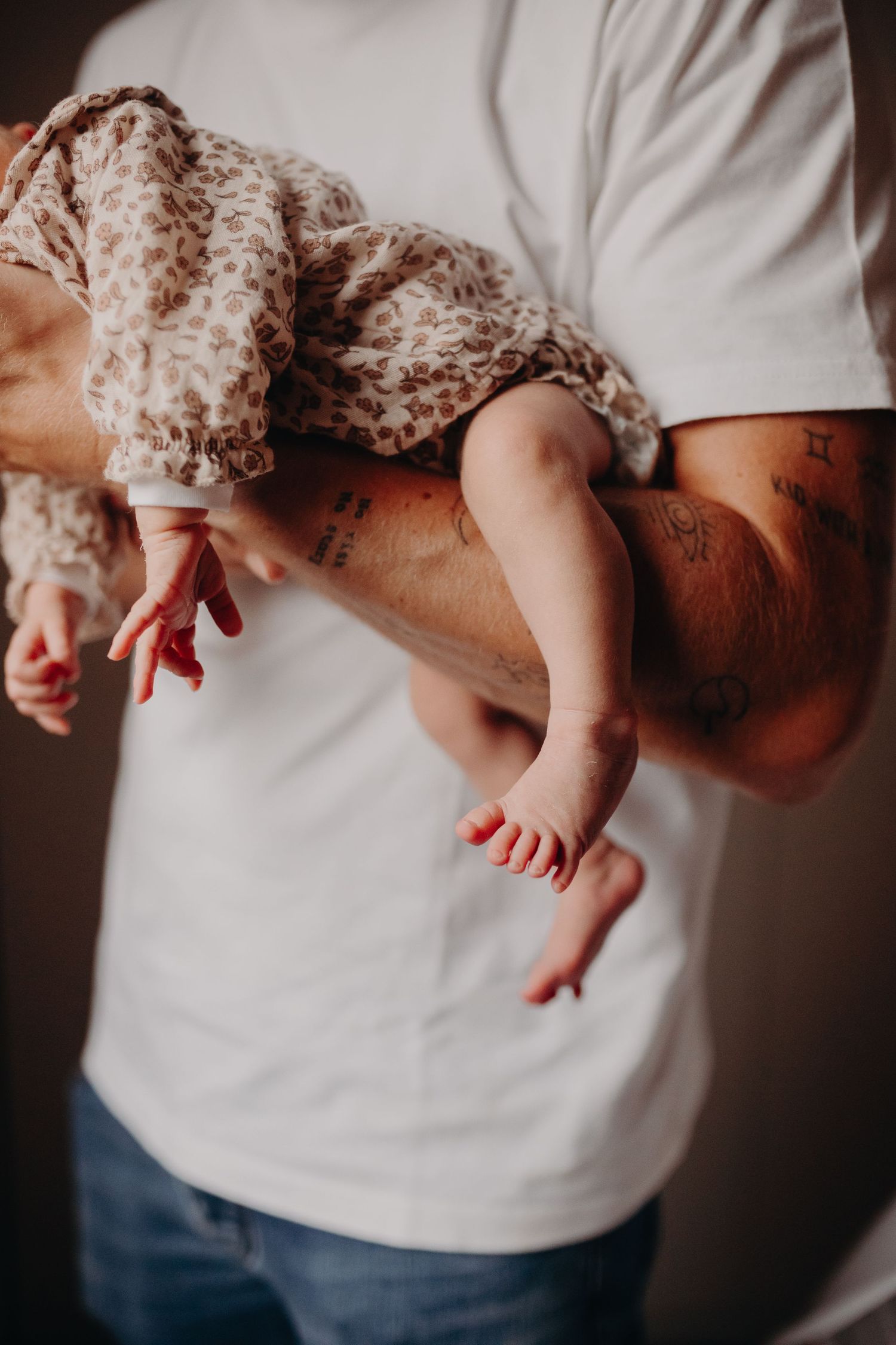 Baby in floral dress being held up showing tiny legs and feet against white shirt and denim jeans background.