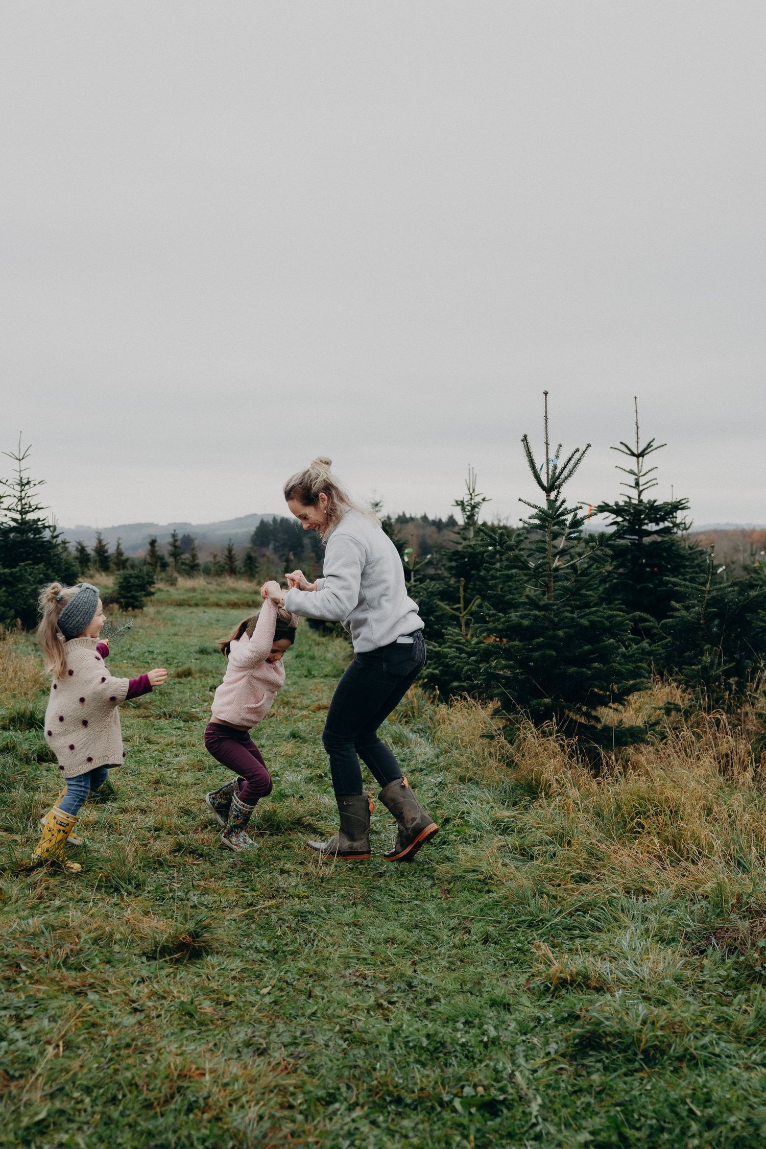 People playing and running together on a grassy hillside with evergreen trees in the background on an overcast day.