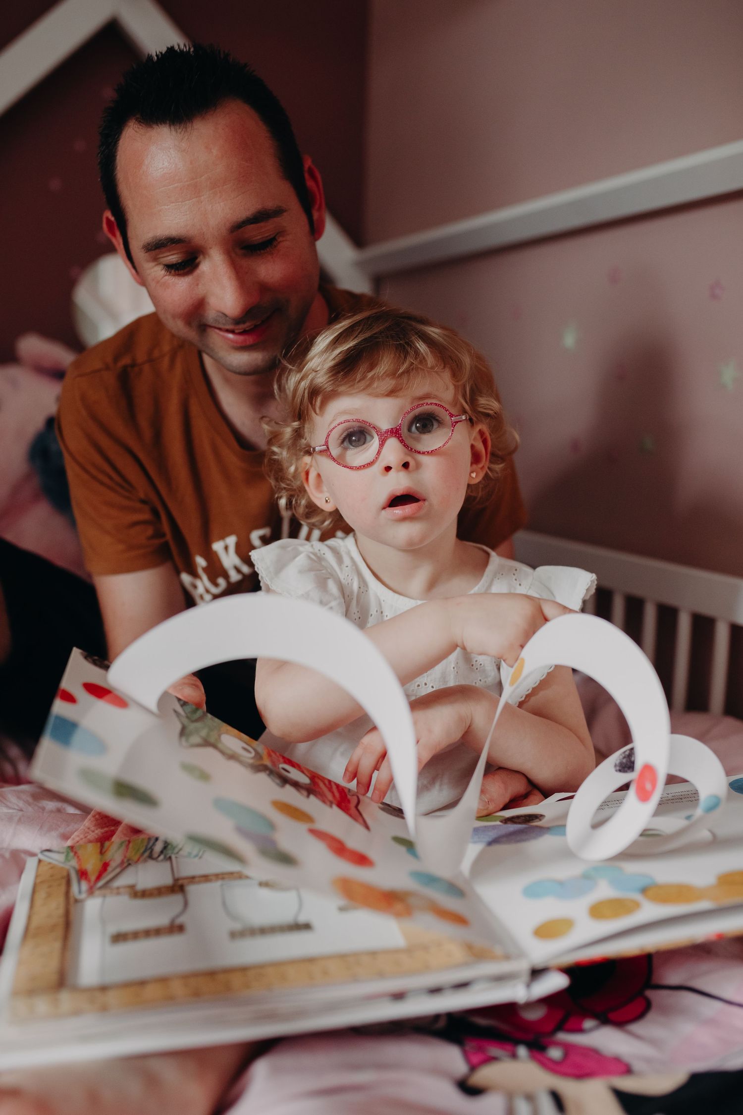 A toddler in round glasses sits with an open picture book while being read to in a cozy bedroom setting.