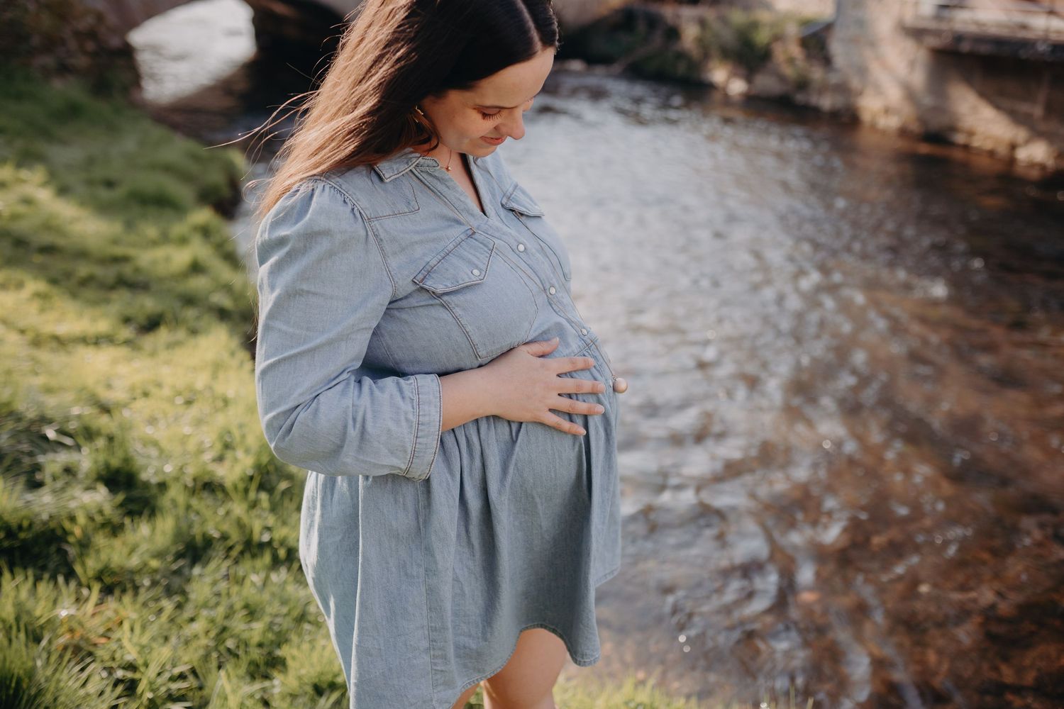 Pregnant woman in blue dress gently cradles her baby bump while standing near a riverbank on a sunny day.