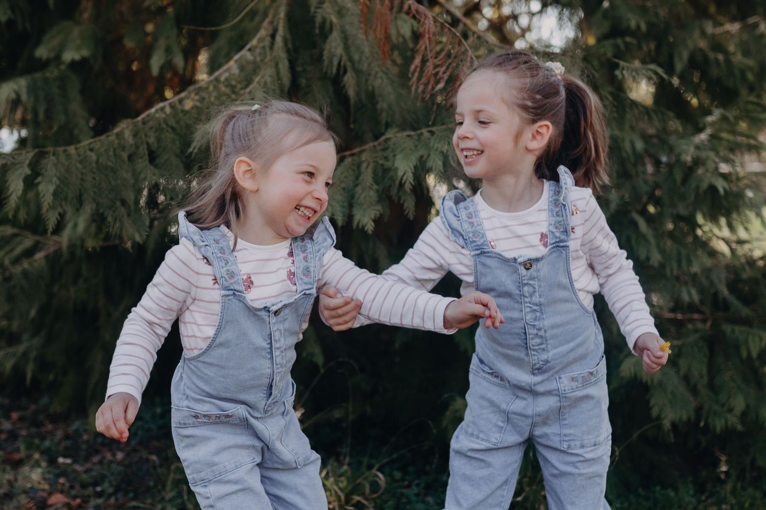 Two children in matching denim overalls and white shirts play and laugh together in front of evergreen trees.