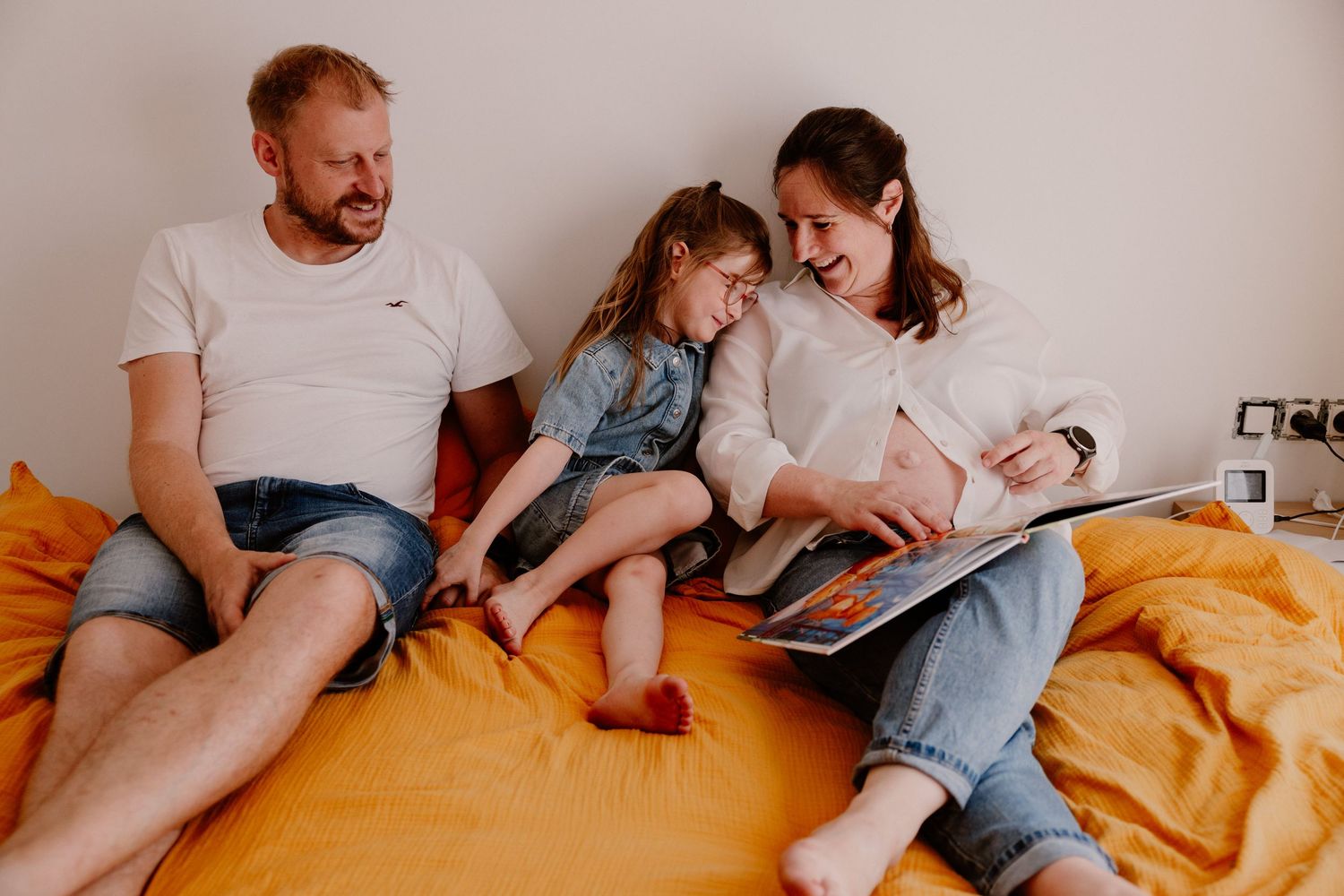 Family of three sitting on bed with orange bedding, parents laughing while reading a picture book with their young daughter.