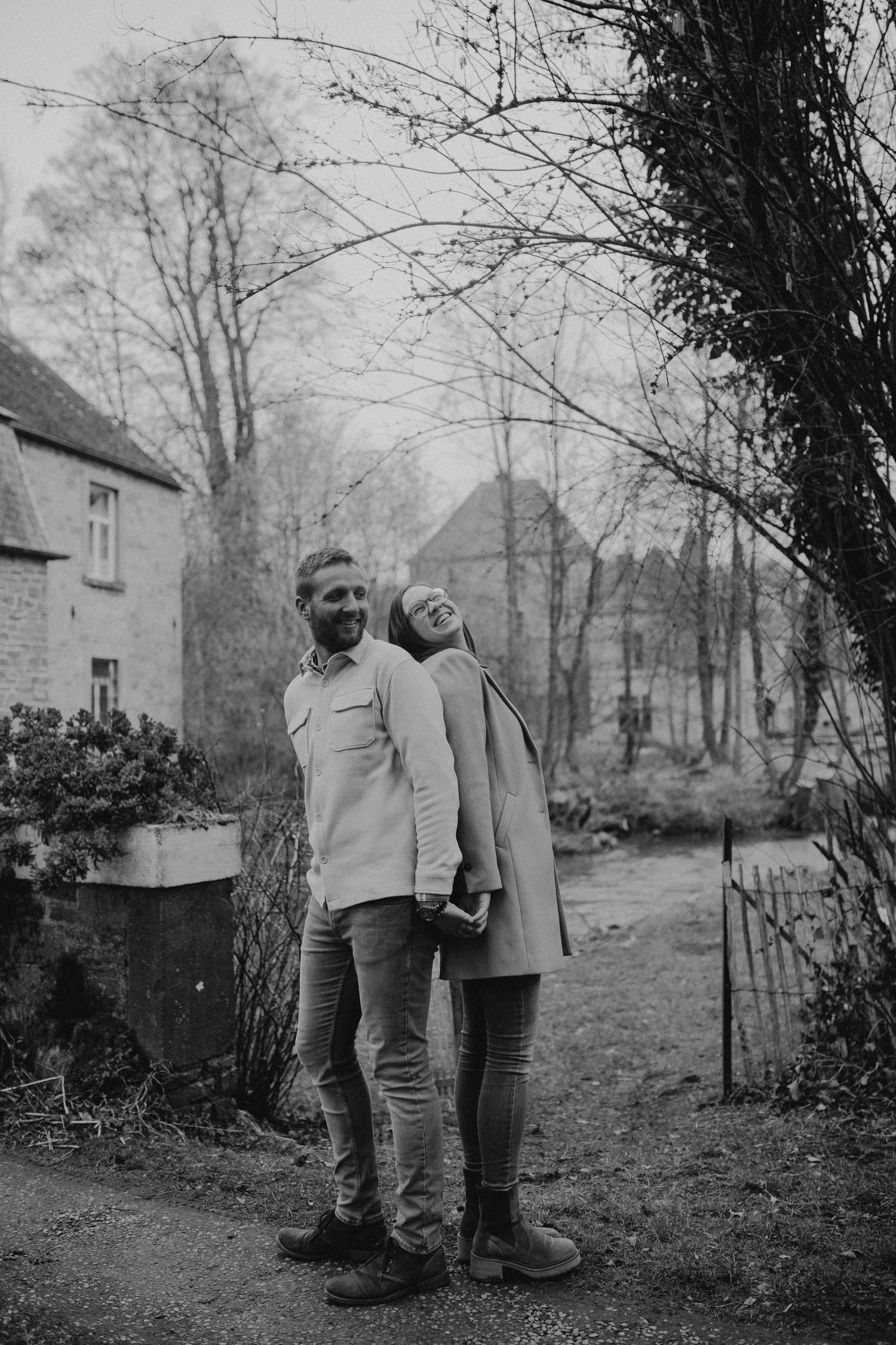 A couple stands together on a garden path in winter with rustic stone buildings in a black and white photo.