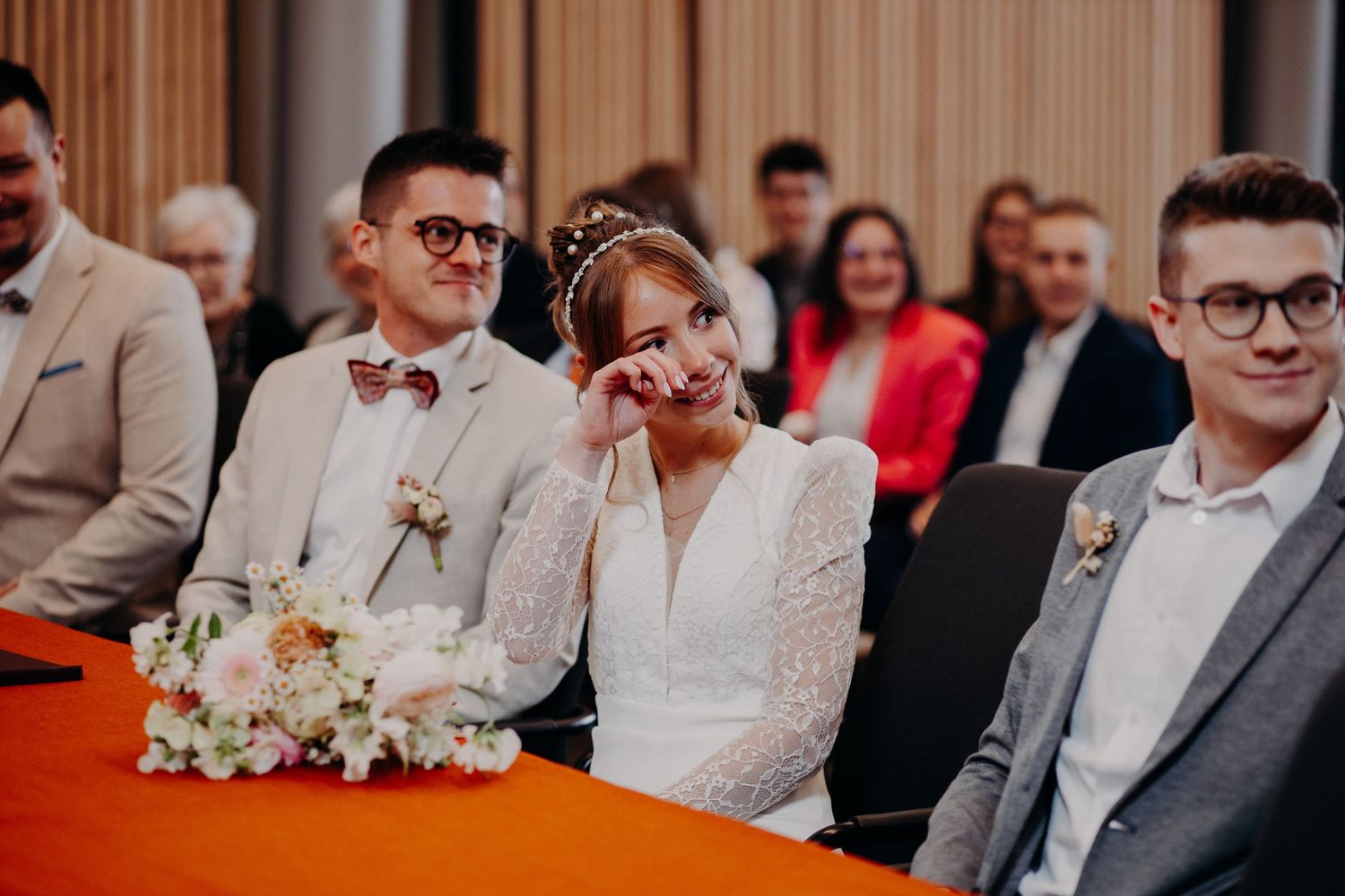 A couple sitting at a wedding ceremony with guests in attendance and floral bouquet details.