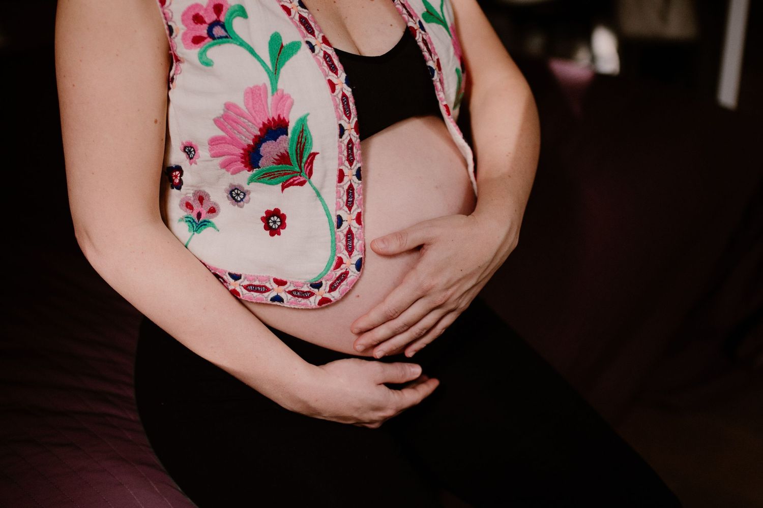 Close-up of pregnant belly wearing a colorful floral embroidered top against dark background.