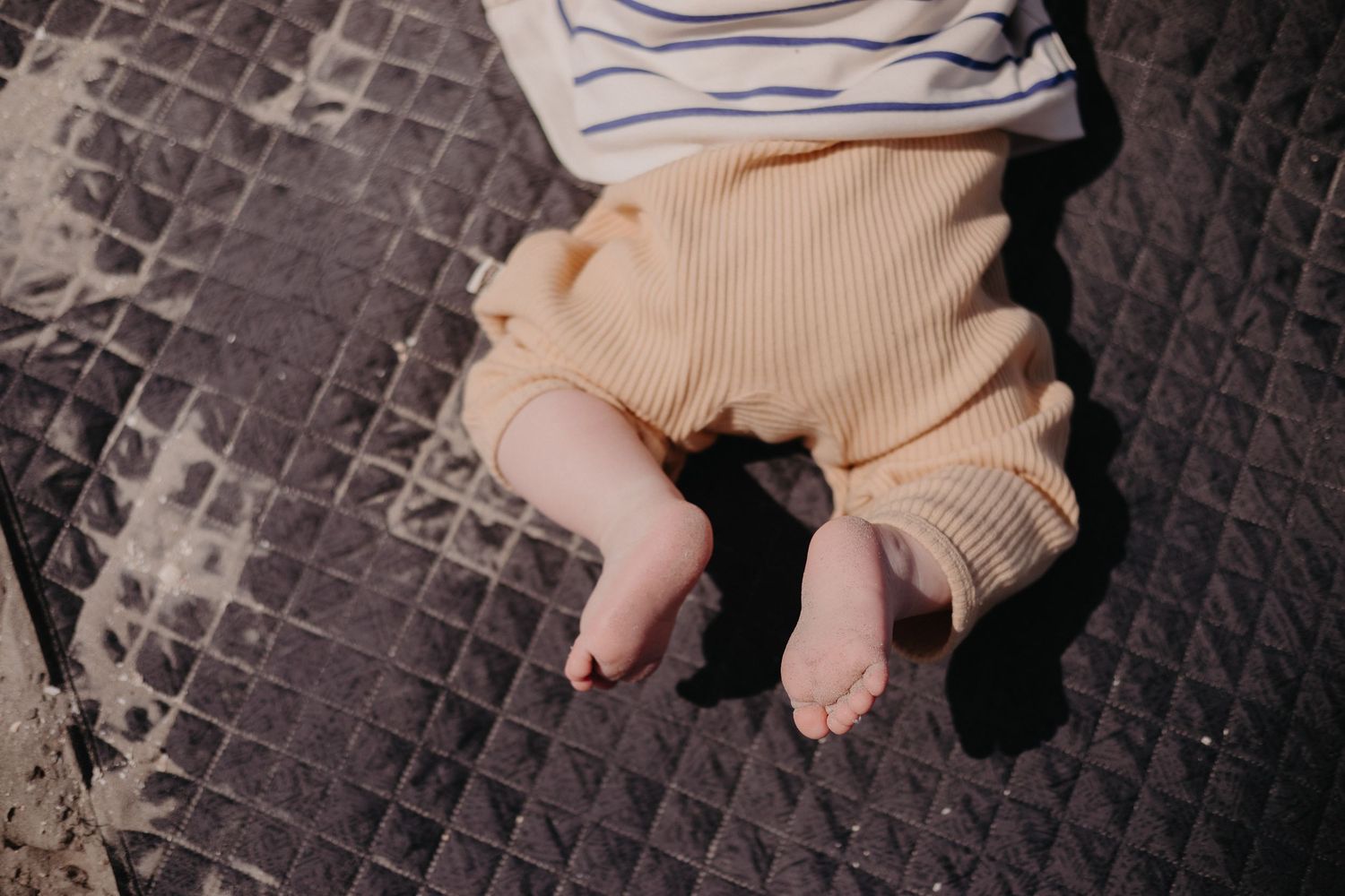 Small bare feet of a baby wearing cozy knit pants resting on a dark textured blanket.