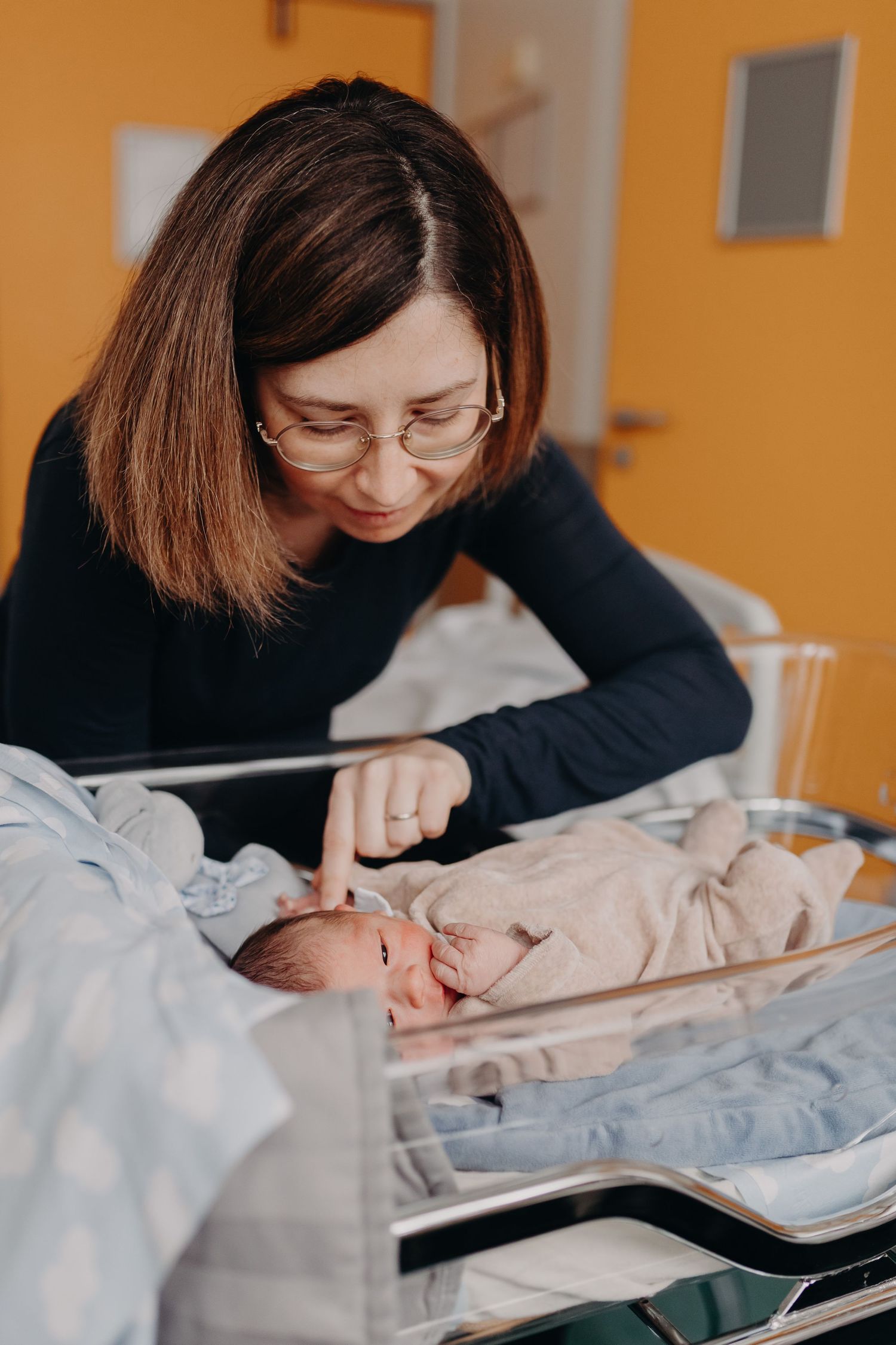 A parent in a black sweater and glasses checks in on a sleeping infant in a medical bassinet.