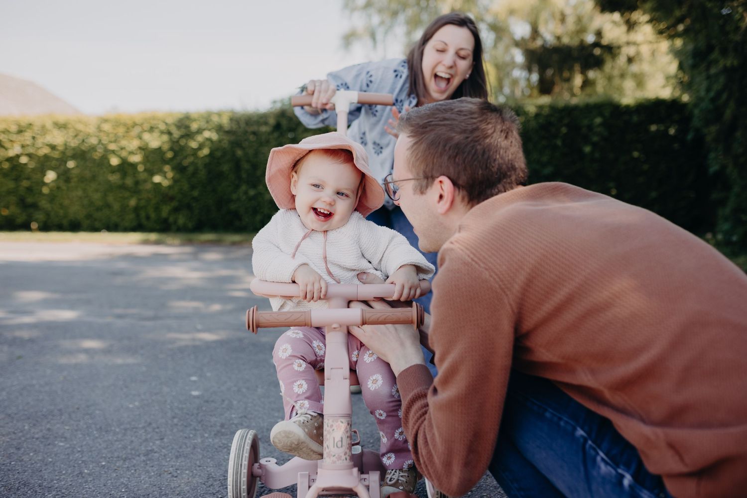 A toddler laughs while sitting in a tricycle on a sunny afternoon with parents on a paved walkway.