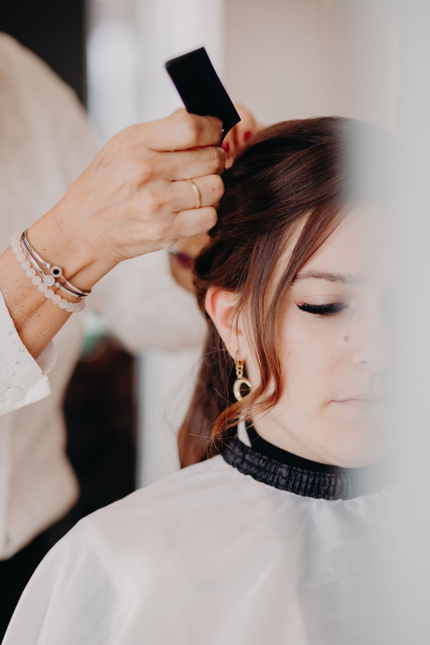 Professional hairstylist styling client's hair in salon while wearing white protective cape and silver bracelets.