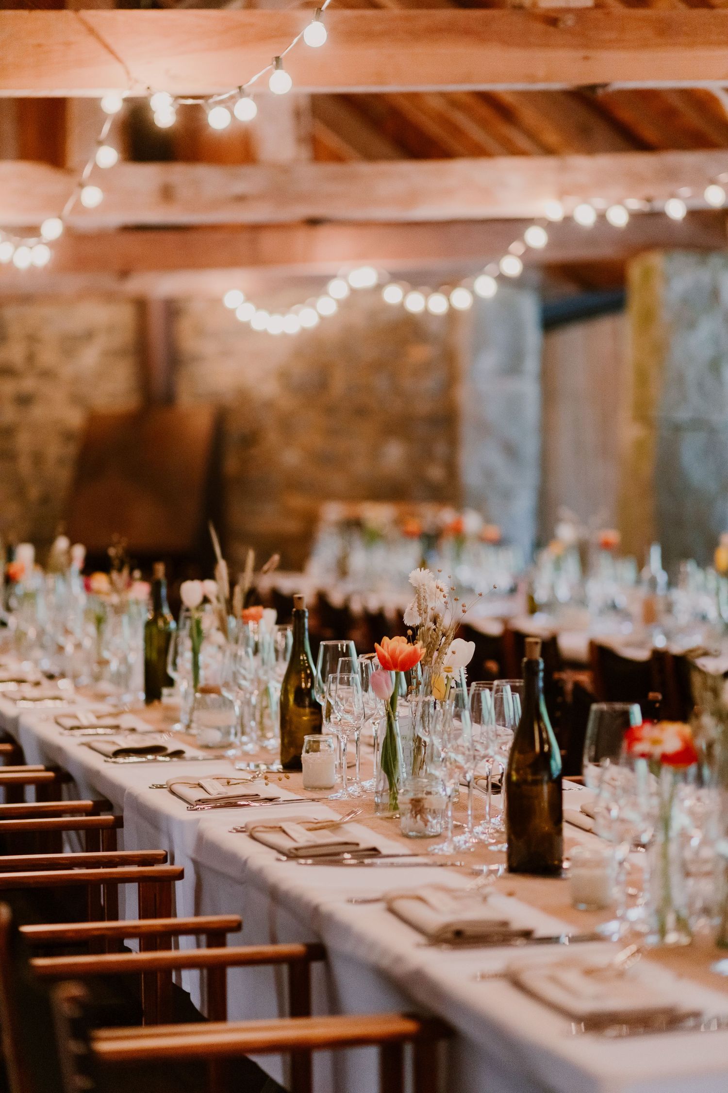 Rustic wedding reception table setting with string lights overhead and wine bottles in a barn venue with stone walls.