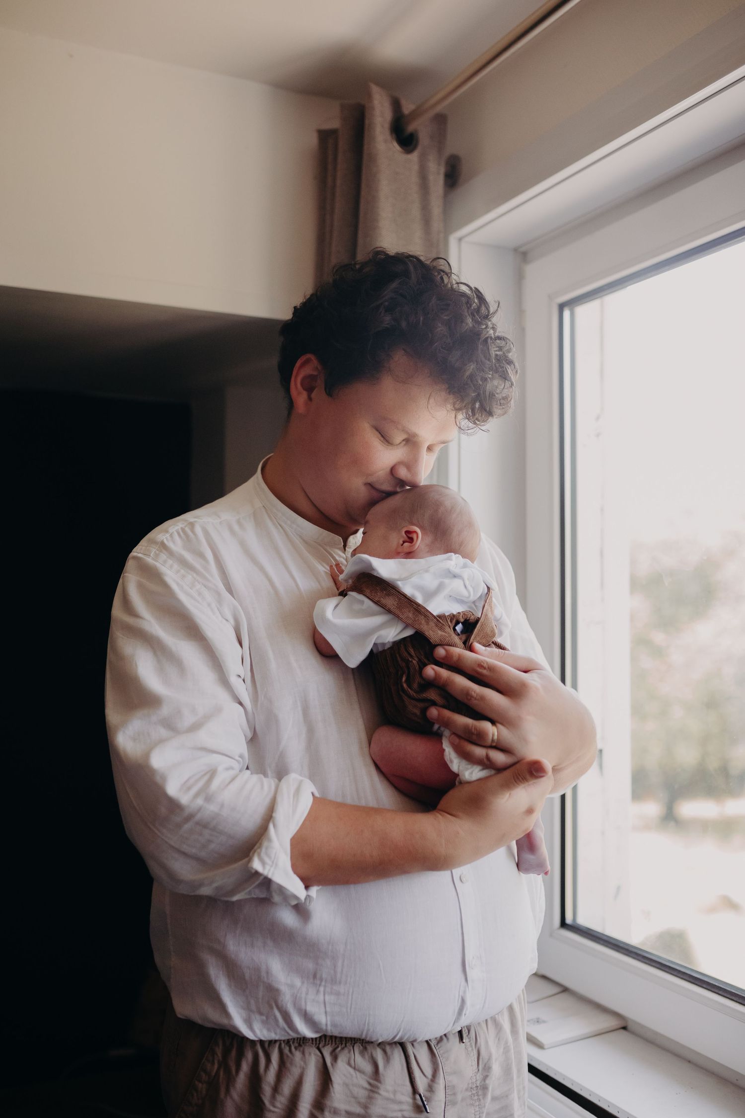 A person in a white shirt holds and gazes lovingly at a small infant by a bright window.