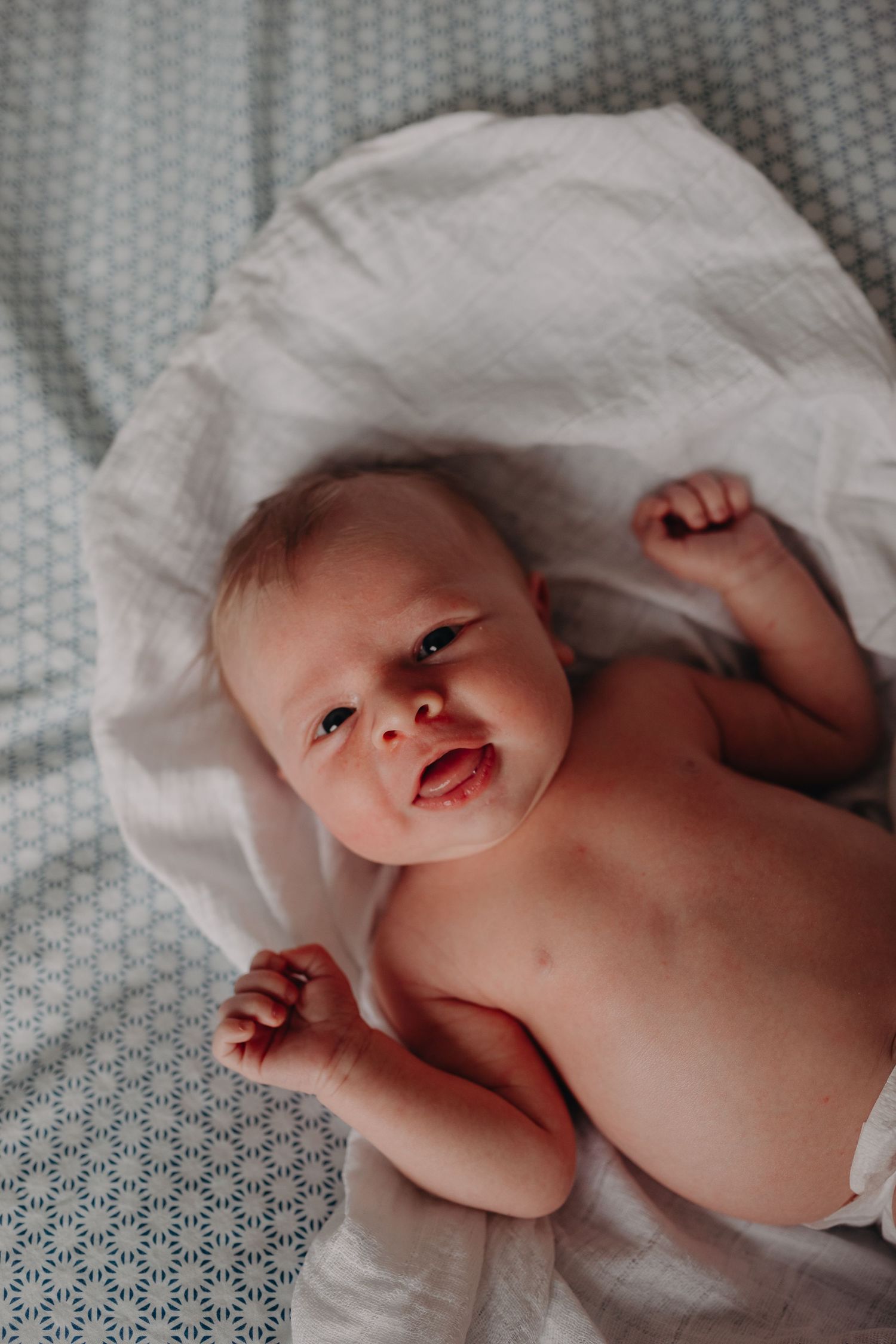 Sweet baby wrapped in soft white blanket smiles while lying on geometric patterned bedding.