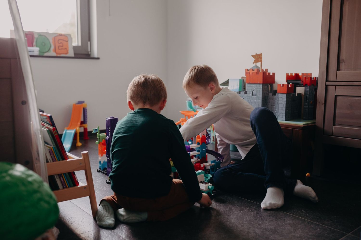 Two children playing with toys together on the floor of a bedroom near a window and dark furniture.