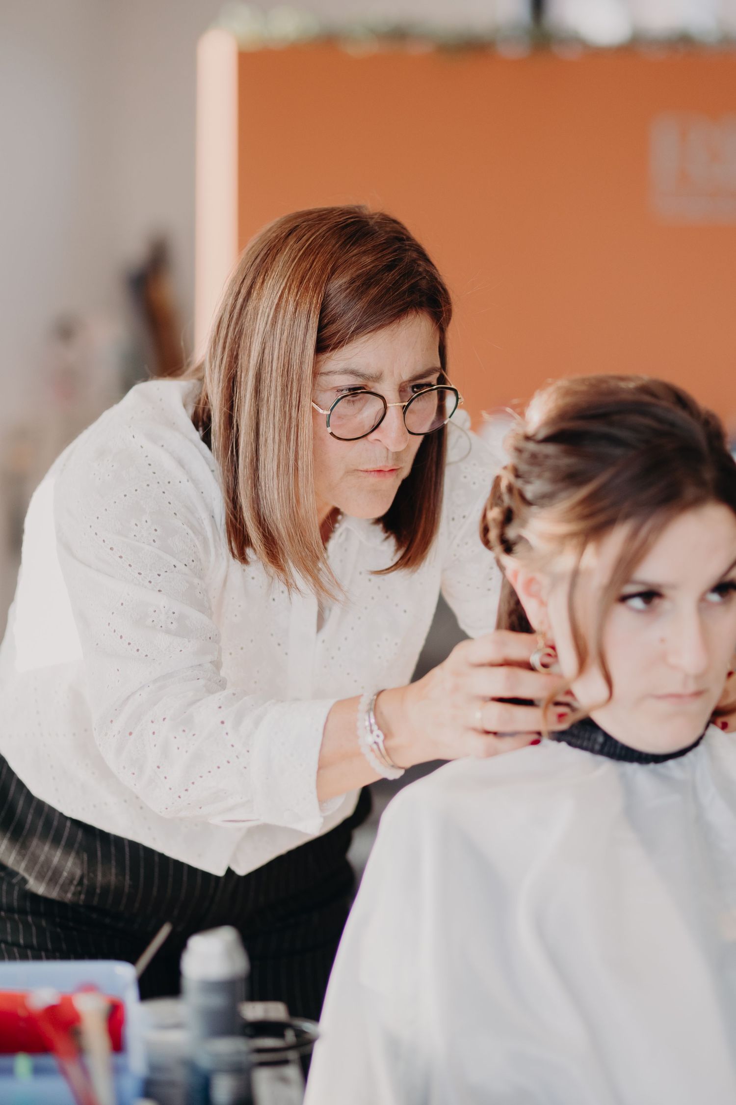 A hairstylist wearing glasses and a white sweater styles a client's hair in a salon setting.