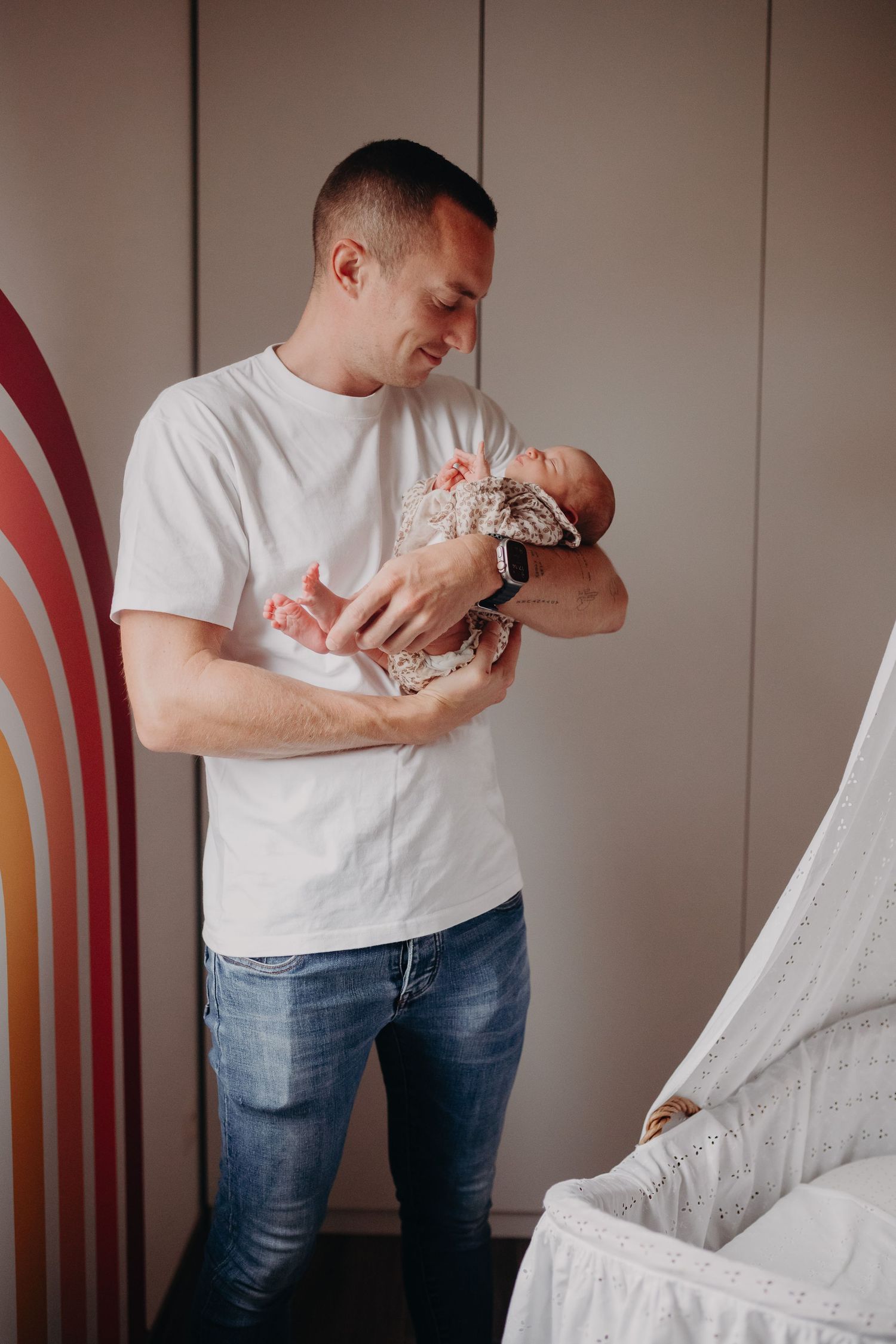 A gentle moment captured as someone in a white t-shirt cradles a newborn baby against a rainbow wall backdrop.